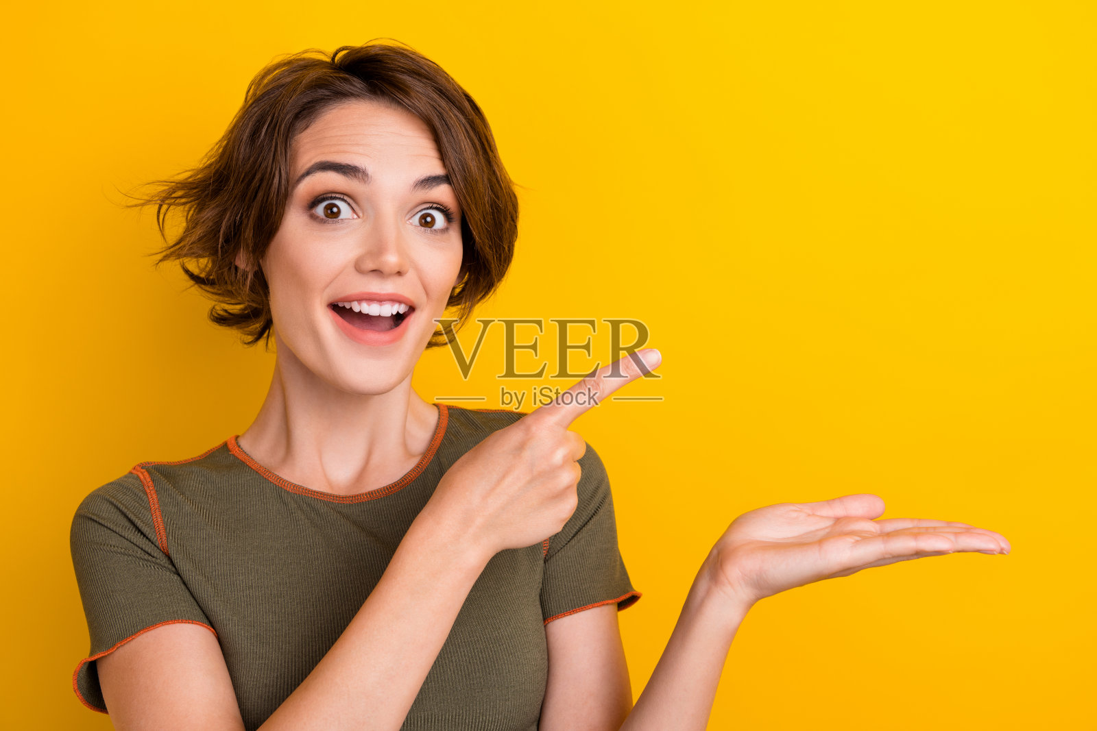 照片的无言欣喜若狂的女孩穿着卡其布t恤指导空的空间手臂持有产品孤立的黄色背景照片摄影图片