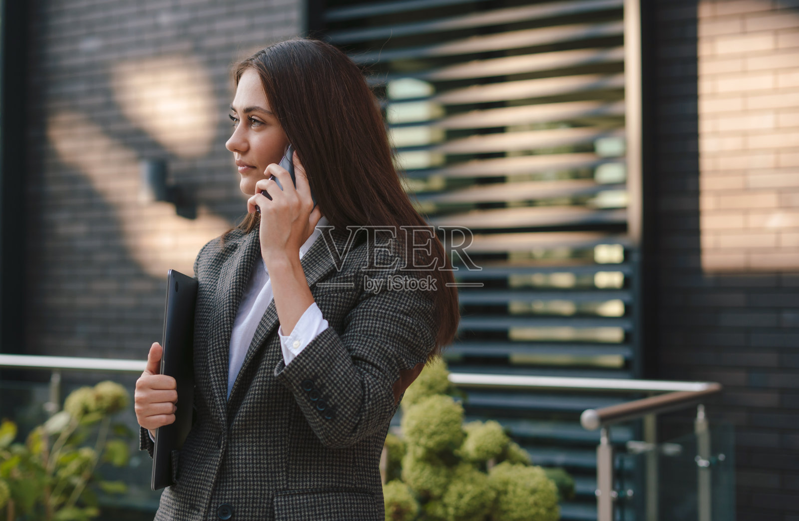 正装的年轻女商人站在现代办公大楼前讲手机的侧面图照片摄影图片