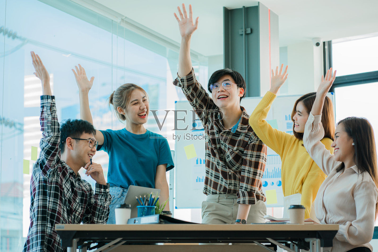年轻的亚洲商业团体感到快乐和成功。笑着举起双手庆祝工作目标的成就,处理桌上的笔记本电脑和办公室的后台财务图。照片摄影图片