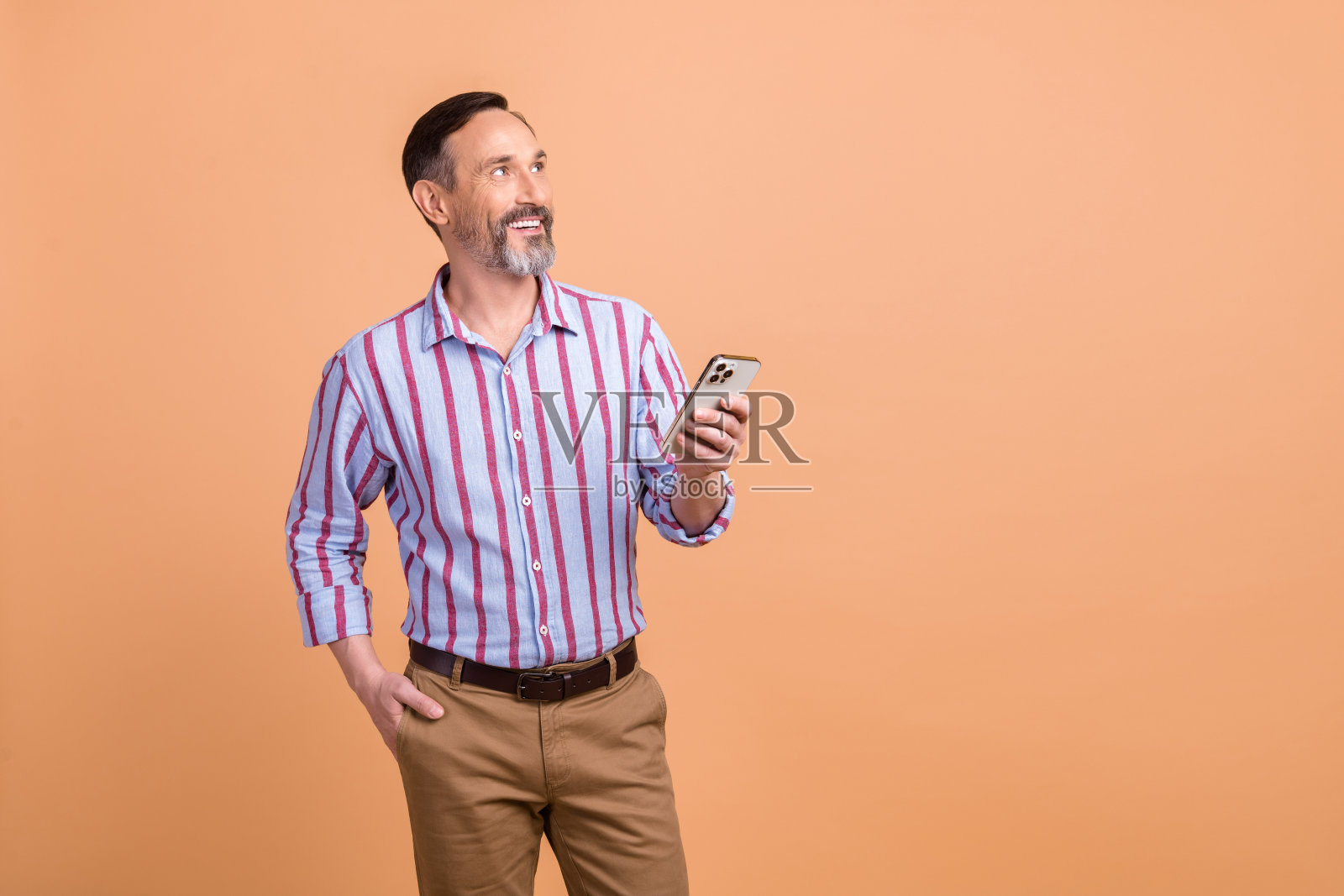 照片中愉快友好的男人穿着条纹衬衫棕色裤子,看着空白的空间,拿着智能手机孤立在米色背景上照片摄影图片