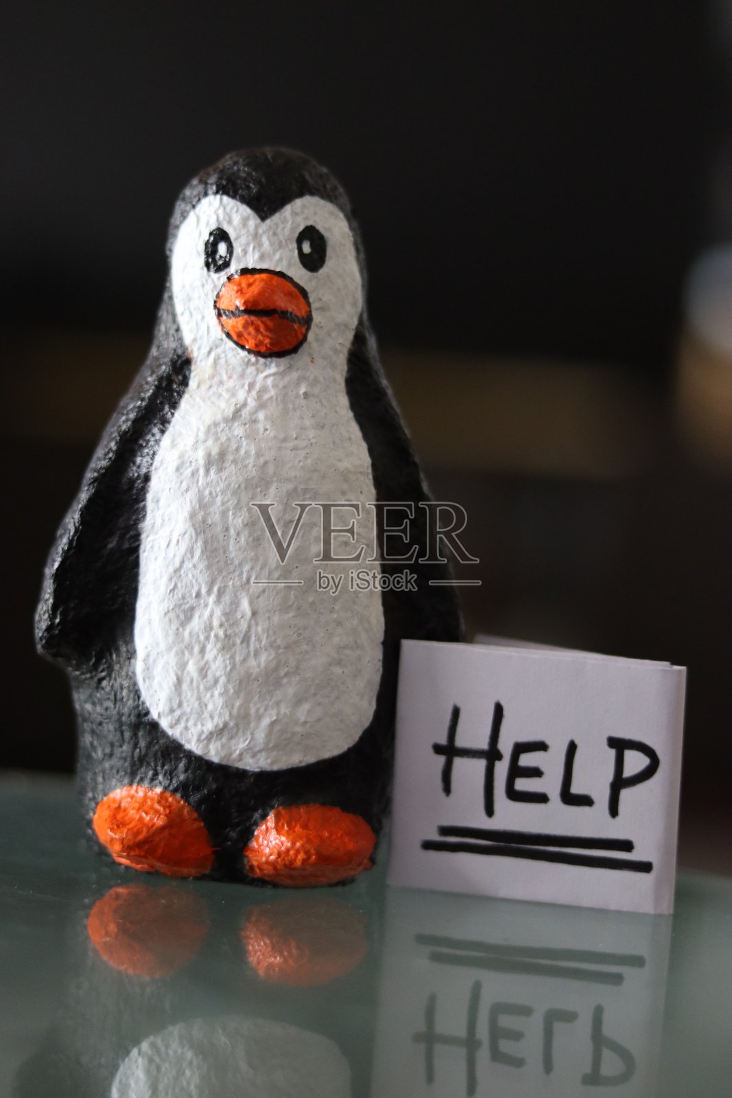 一只用纸浆做的小企鹅,手里拿着一张纸做的“帮助的标志”,旁边有散景照片摄影图片