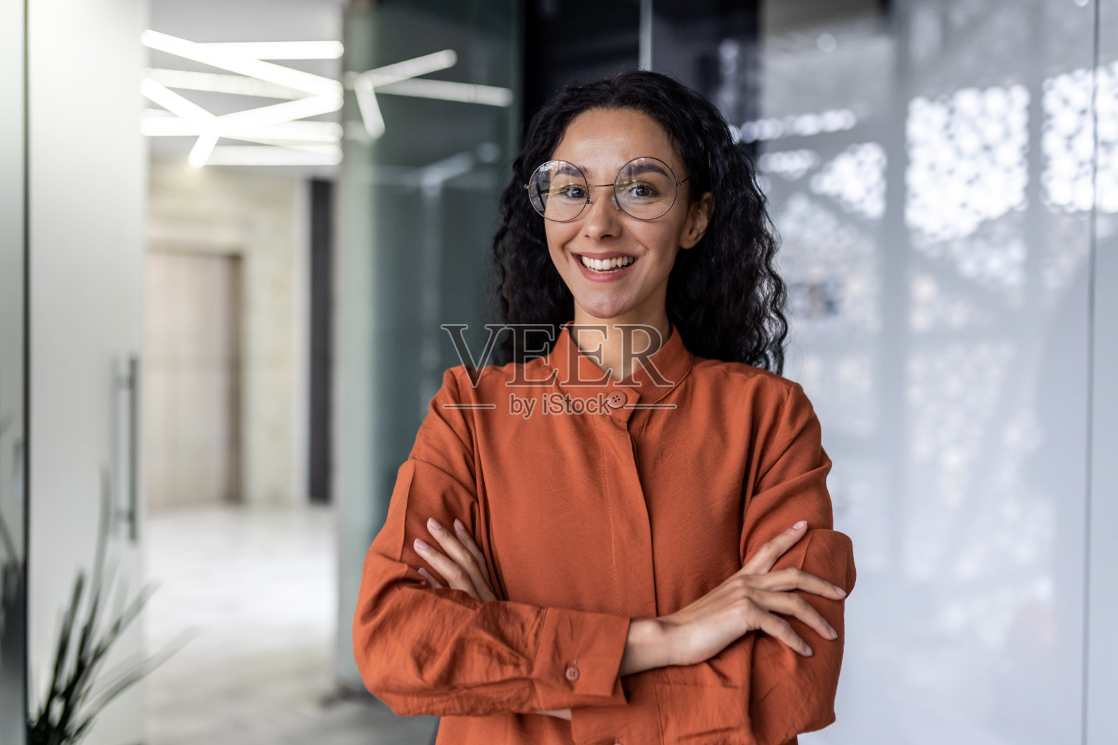年轻美丽的成功西班牙裔女商人微笑地看着镜头,女工在办公室里戴着眼镜和卷发工作,双臂交叉的肖像照片摄影图片