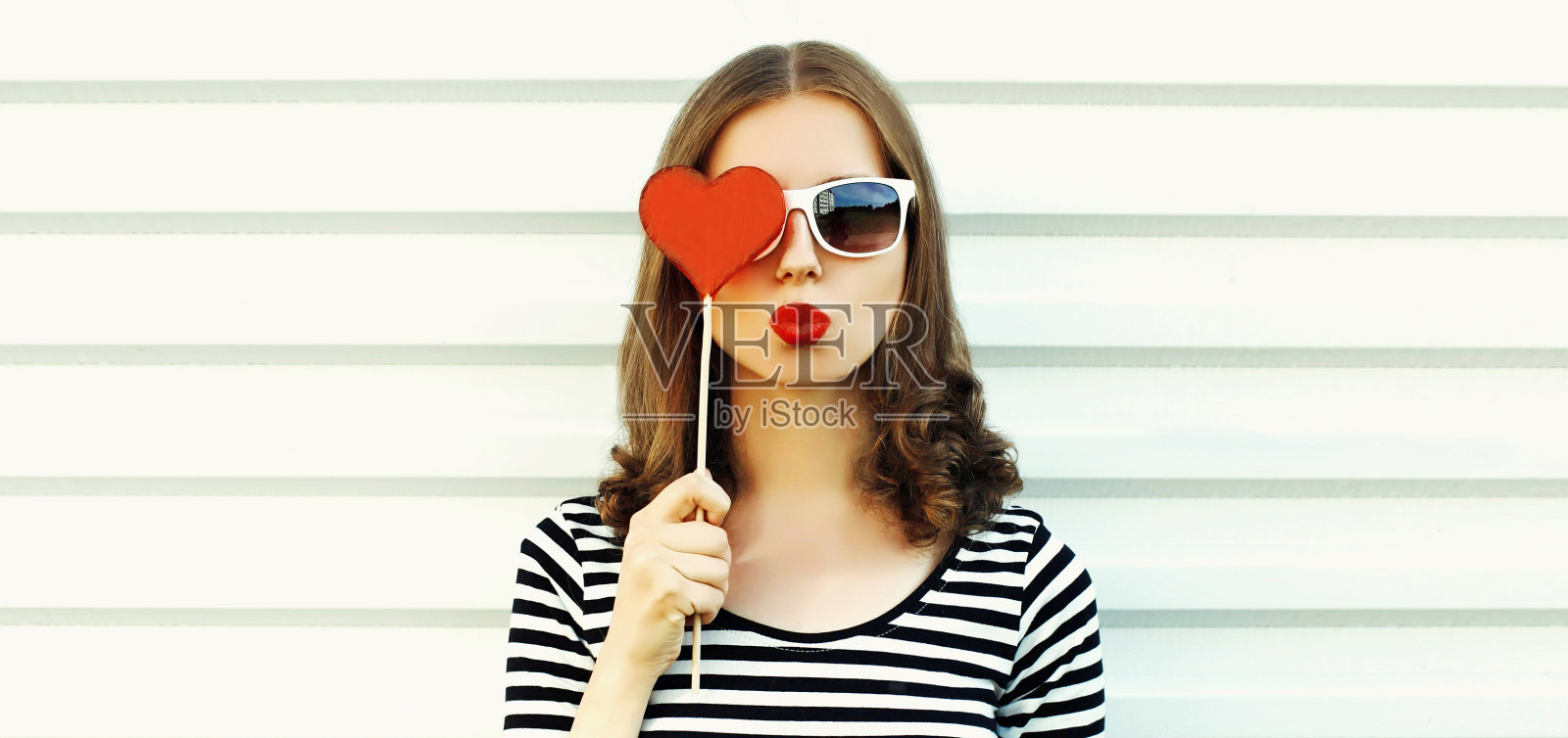 美丽的年轻女子的肖像吹她的嘴唇与红色的甜心形状的棒棒糖口红在白色的背景照片摄影图片