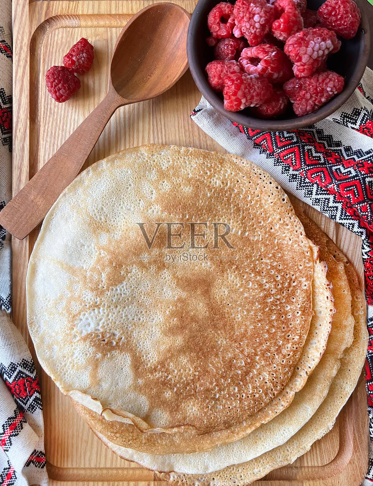 美味可口的早餐,可丽饼和覆盆子。忏悔节假期。Maslenitsa派对——社交活动。用勺子和刺绣衬衫在木板上做煎饼。俯视图照片摄影图片