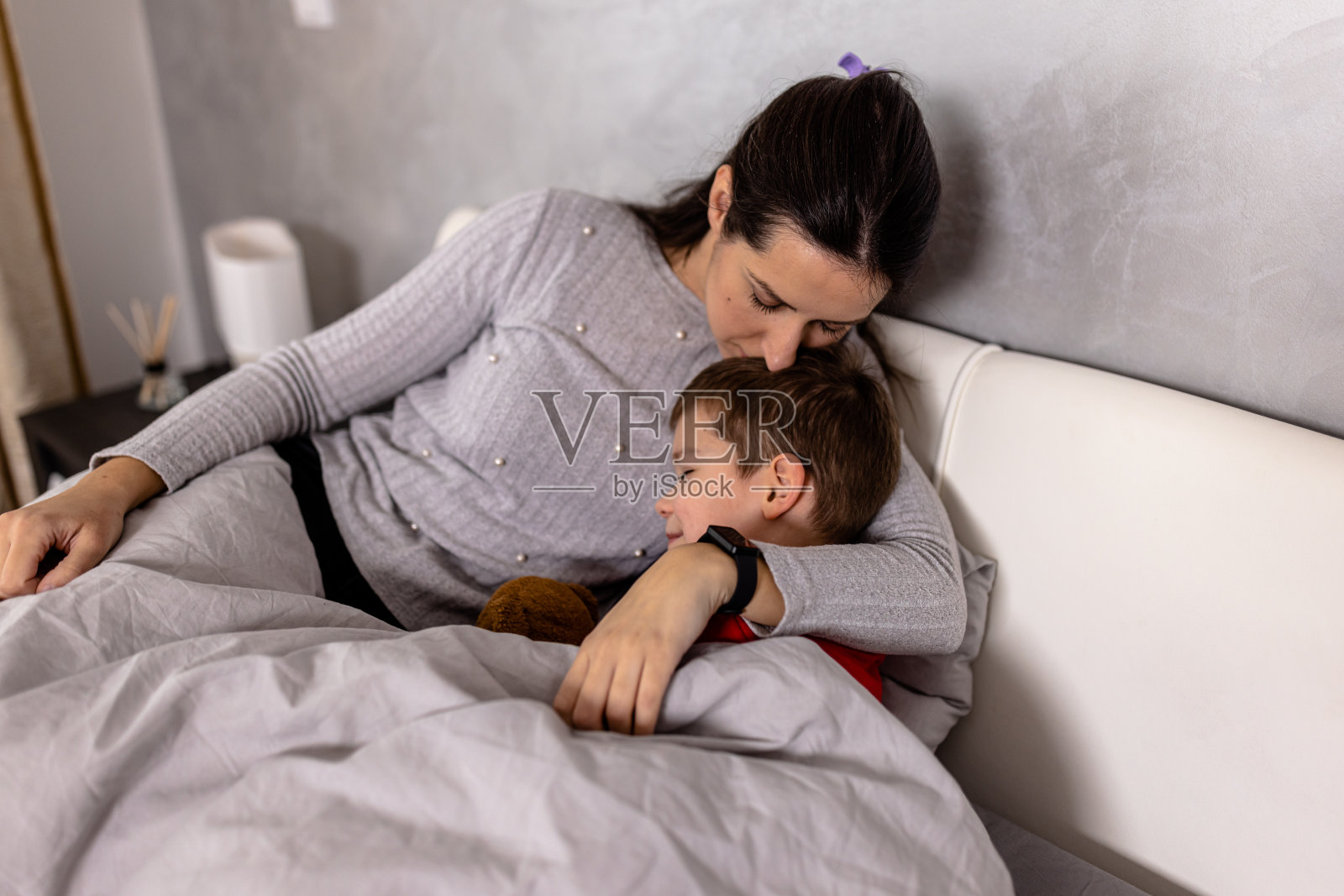 一位年轻的母亲抱着儿子躺在卧室里,拥抱着他。照片摄影图片