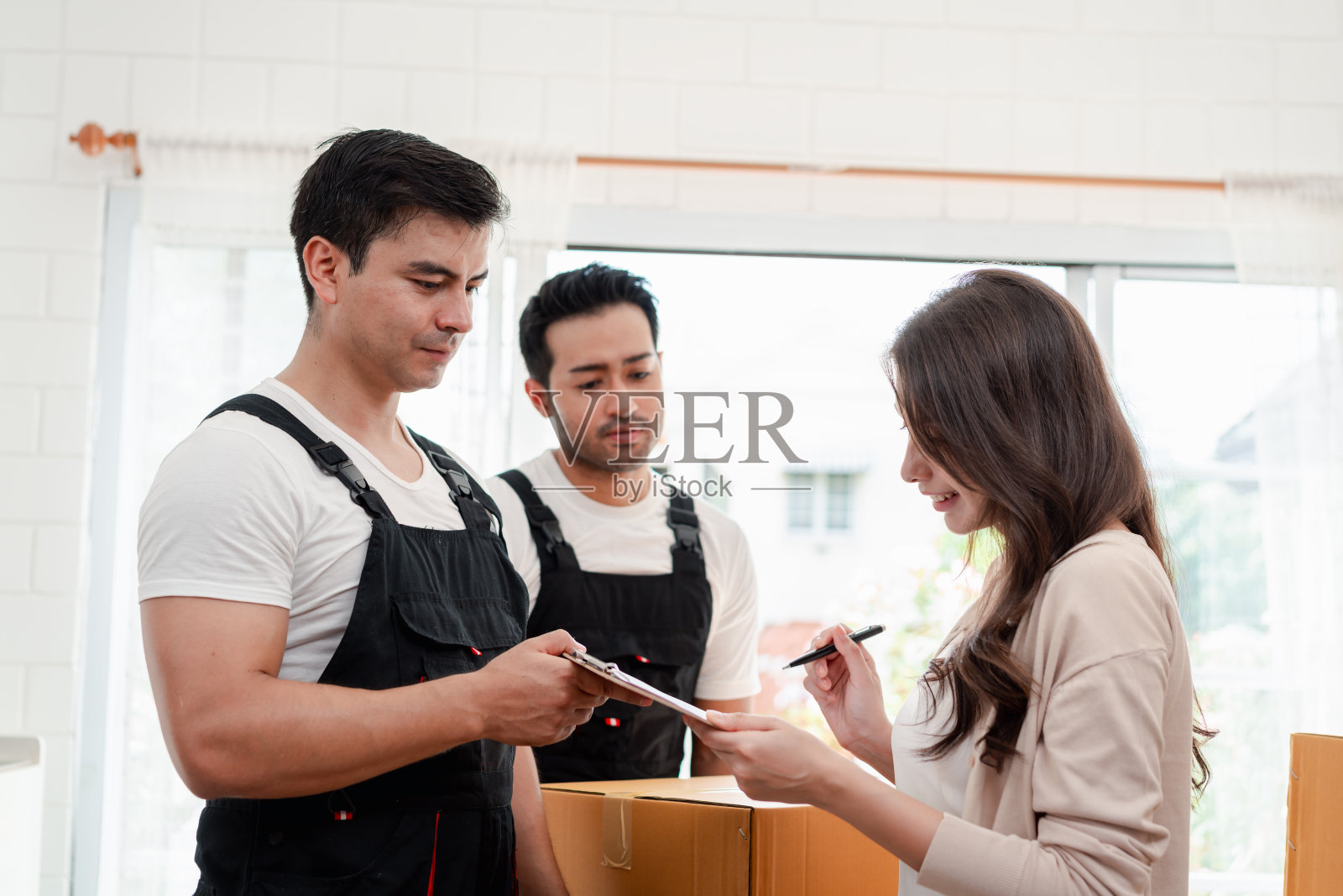 一名妇女一边检查,一边把箱子和家具从一辆小货车上卸到一所有服务货物的新房子里,两名穿着制服的搬运工正在搬运箱子。搬家和送货的概念。照片摄影图片