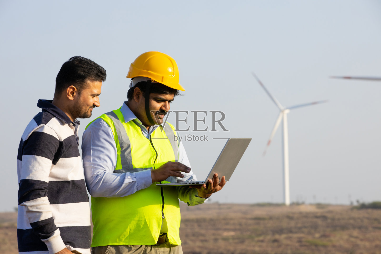 印度工程师使用笔记本电脑,展示控制风车农场发电的操作,亚洲人戴着黄色头盔和背心,在风力涡轮机农场工作,清洁和绿色能源。照片摄影图片