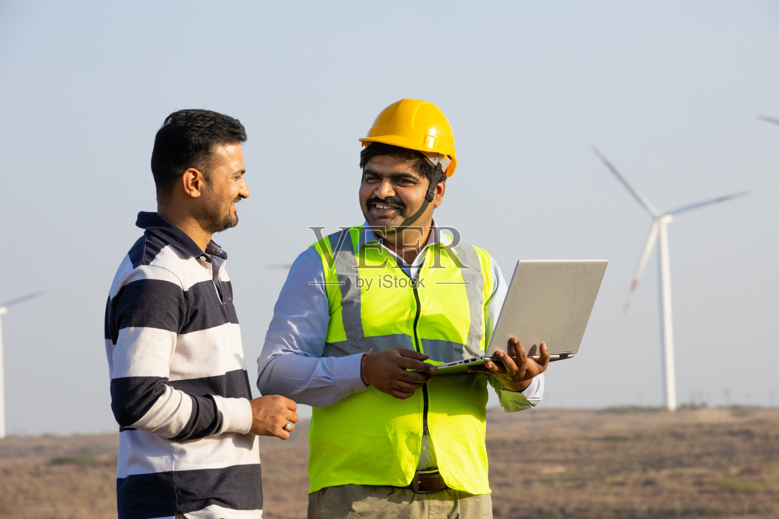 印度工程师使用笔记本电脑,讨论风车农场的运作发电,亚洲人戴着黄色头盔和背心在风力涡轮机农场工作,清洁和绿色能源。照片摄影图片