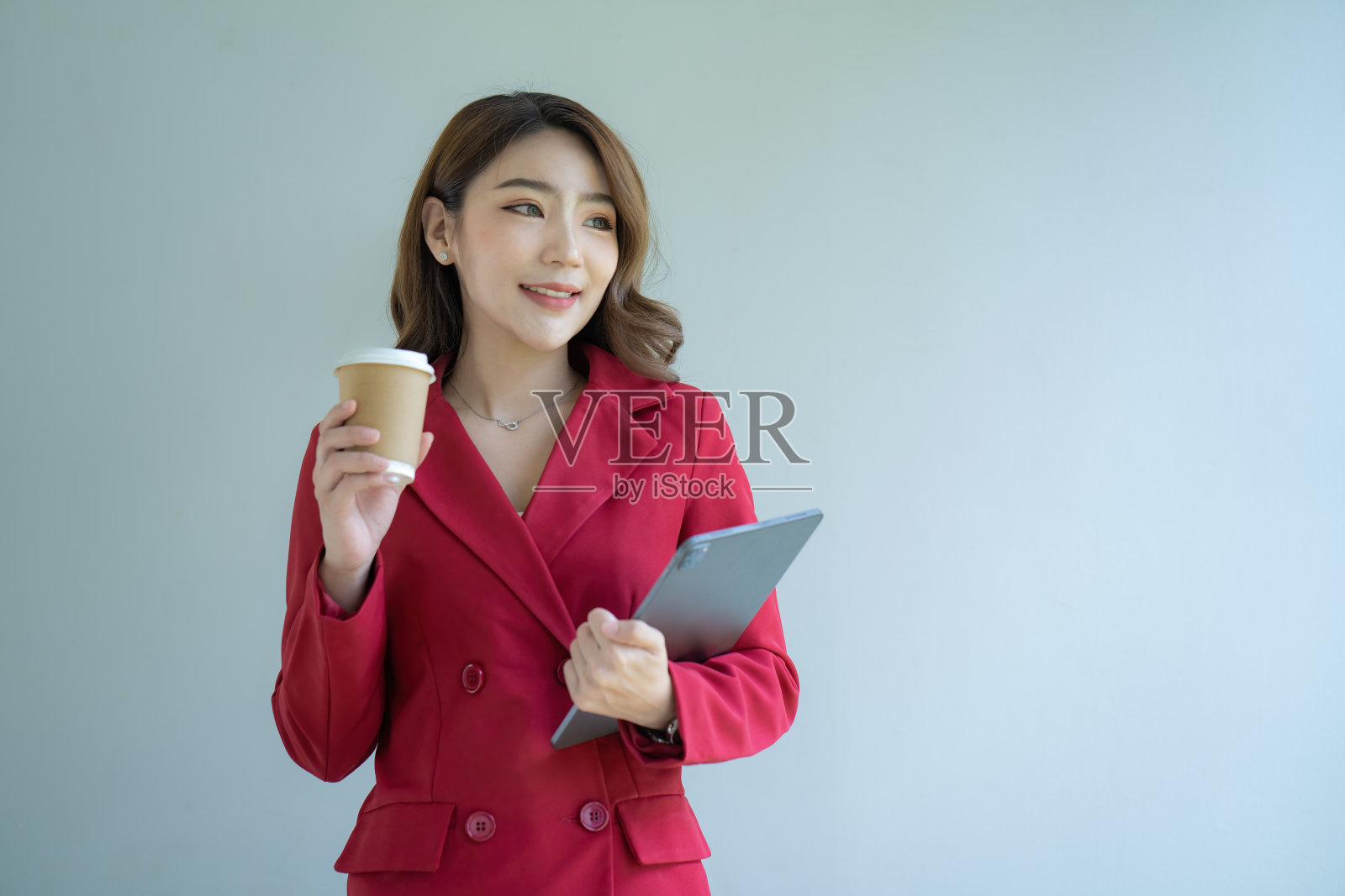 身穿红色西装的亚洲女商人拿着一台数码平板电脑和一杯咖啡,背景是白色。照片摄影图片