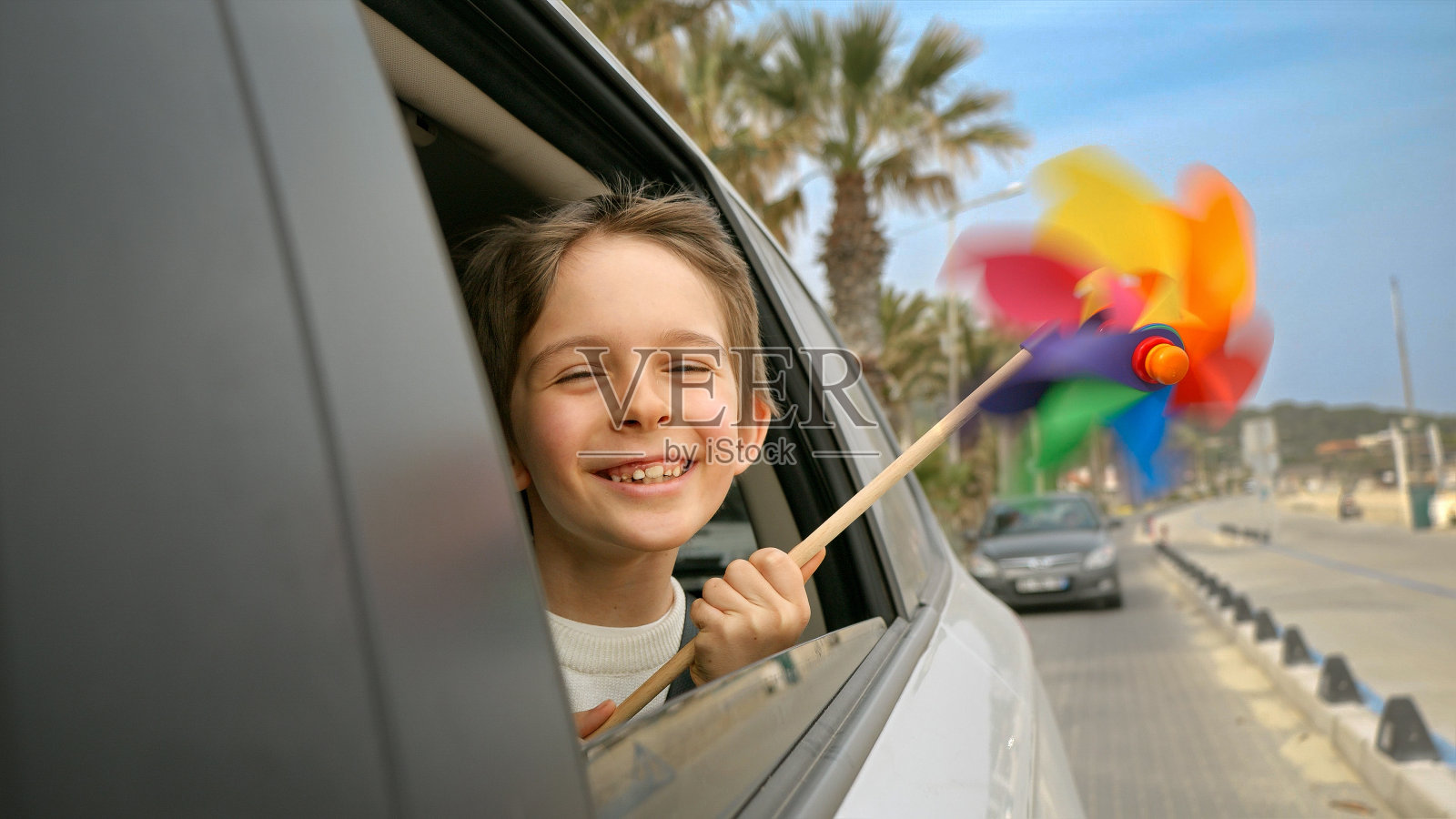 开心微笑的男孩拿着彩色的风向标或风车,看着打开的车窗外。旅游、旅行、旅行、暑假、假期的概念照片摄影图片