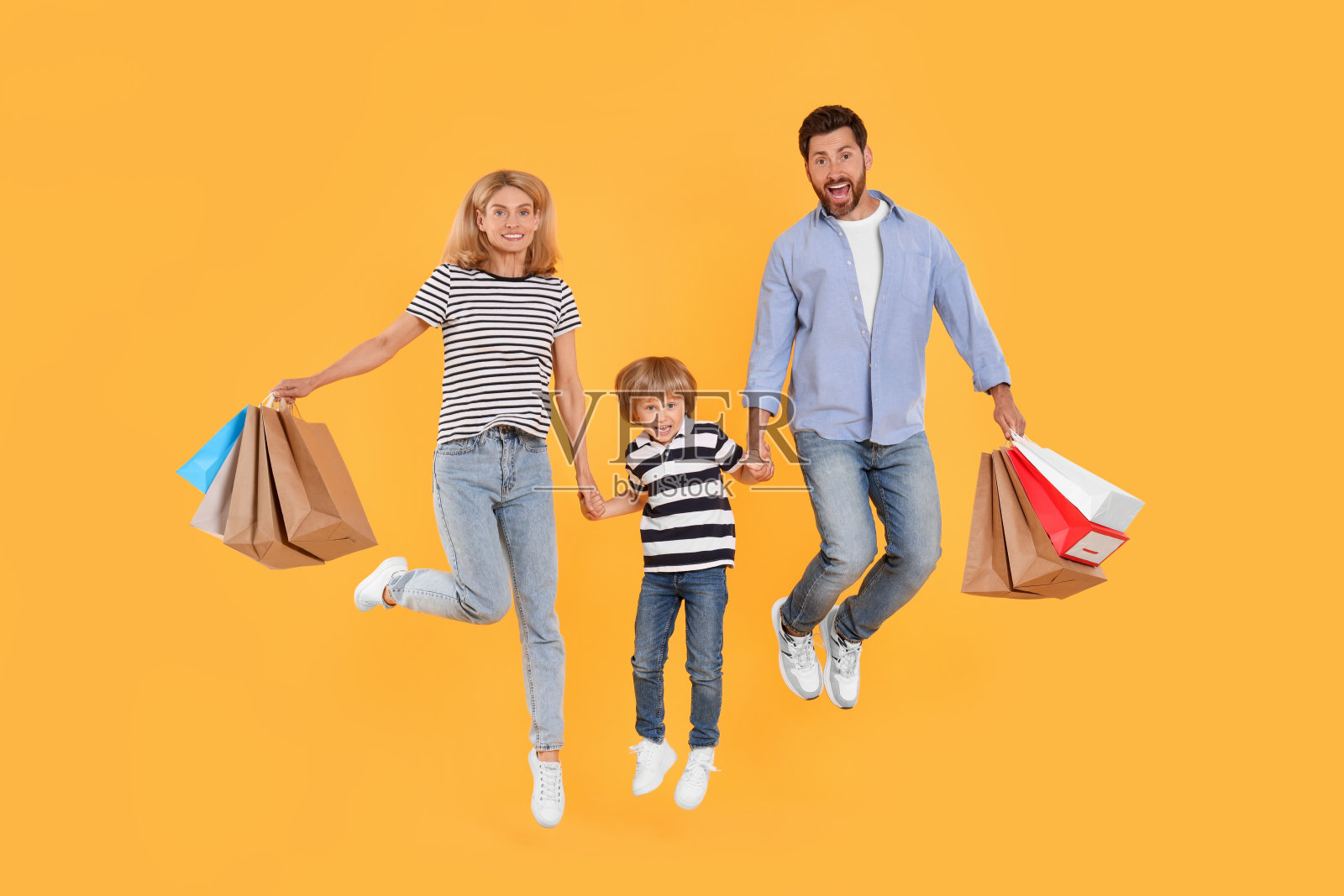 家庭购物。快乐的父母和儿子拿着橘色背景的纸袋蹦蹦跳跳照片摄影图片