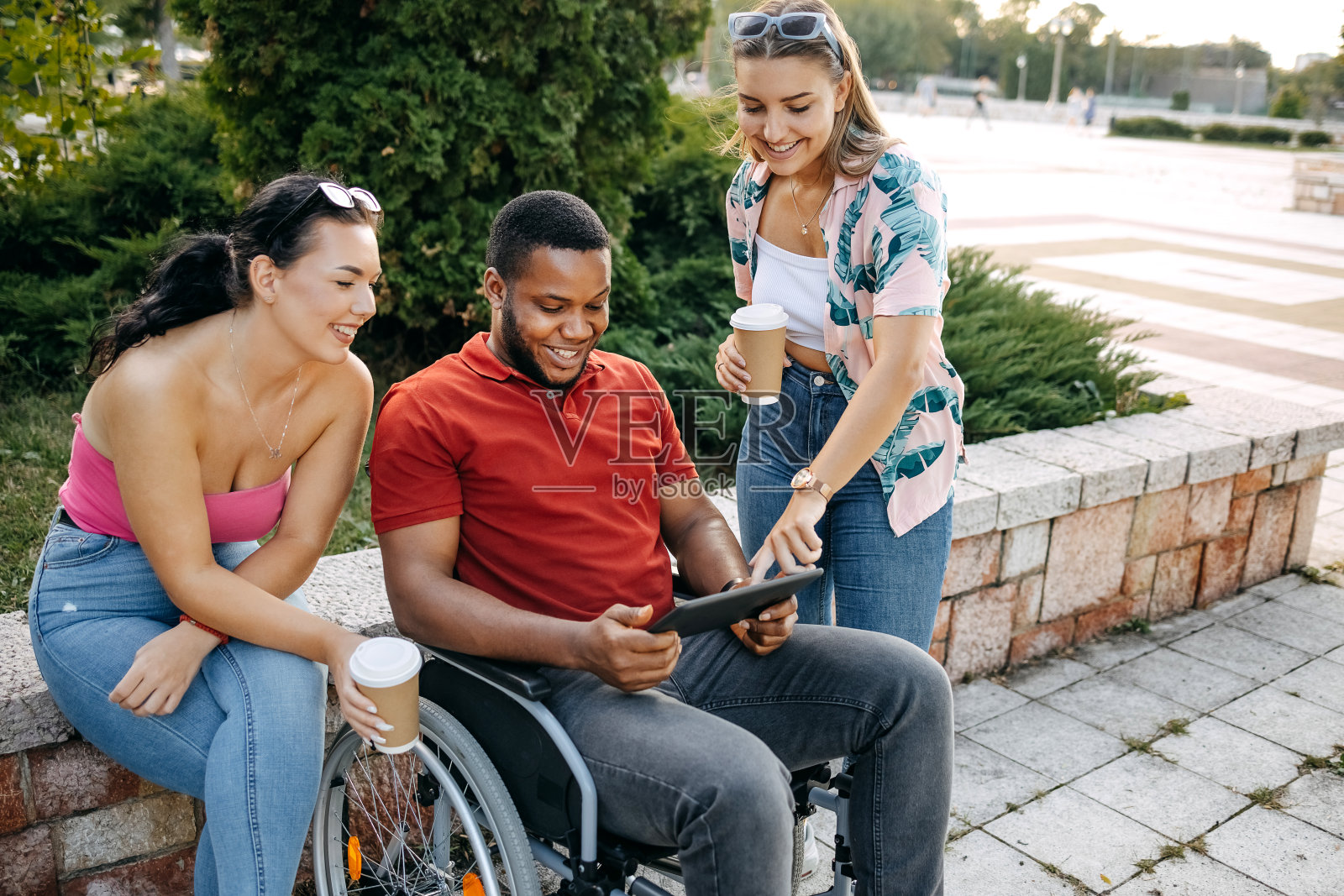 坐在轮椅上的年轻黑人男子和朋友在公园外面玩电子平板电脑照片摄影图片