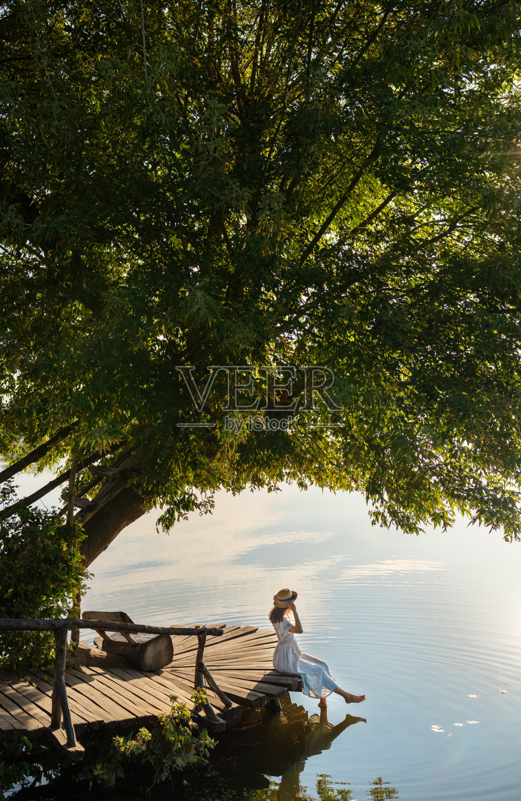 一个年轻漂亮的女孩戴着帽子,穿着长裙,坐在湖边的码头上,看着初升的太阳。旅游和户外娱乐的概念。照片摄影图片