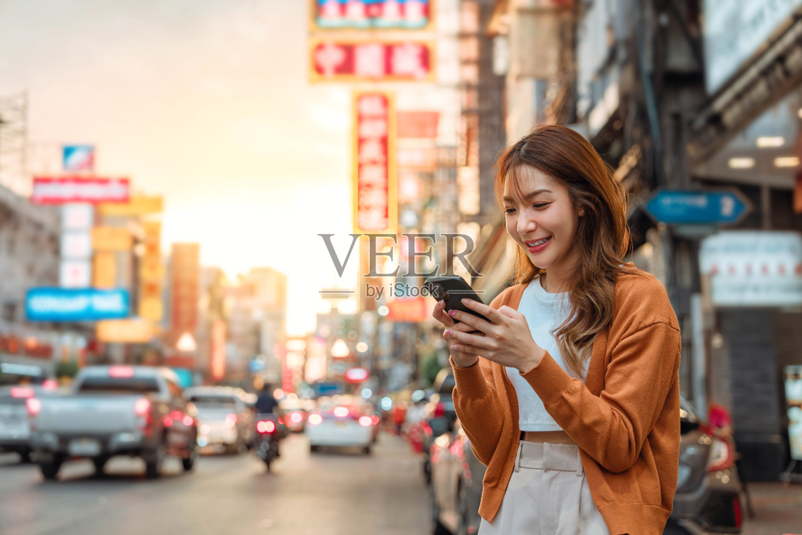 快乐的年轻亚洲女游客在灯光散景背景下的街道上使用智能手机。日落时分,泰国曼谷Yaowarat唐人街街头食品市场的女游客。照片摄影图片