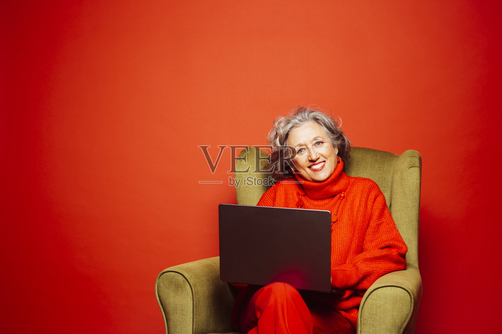 一位年长的女士坐在扶手椅上,用红色背景的笔记本电脑照片摄影图片
