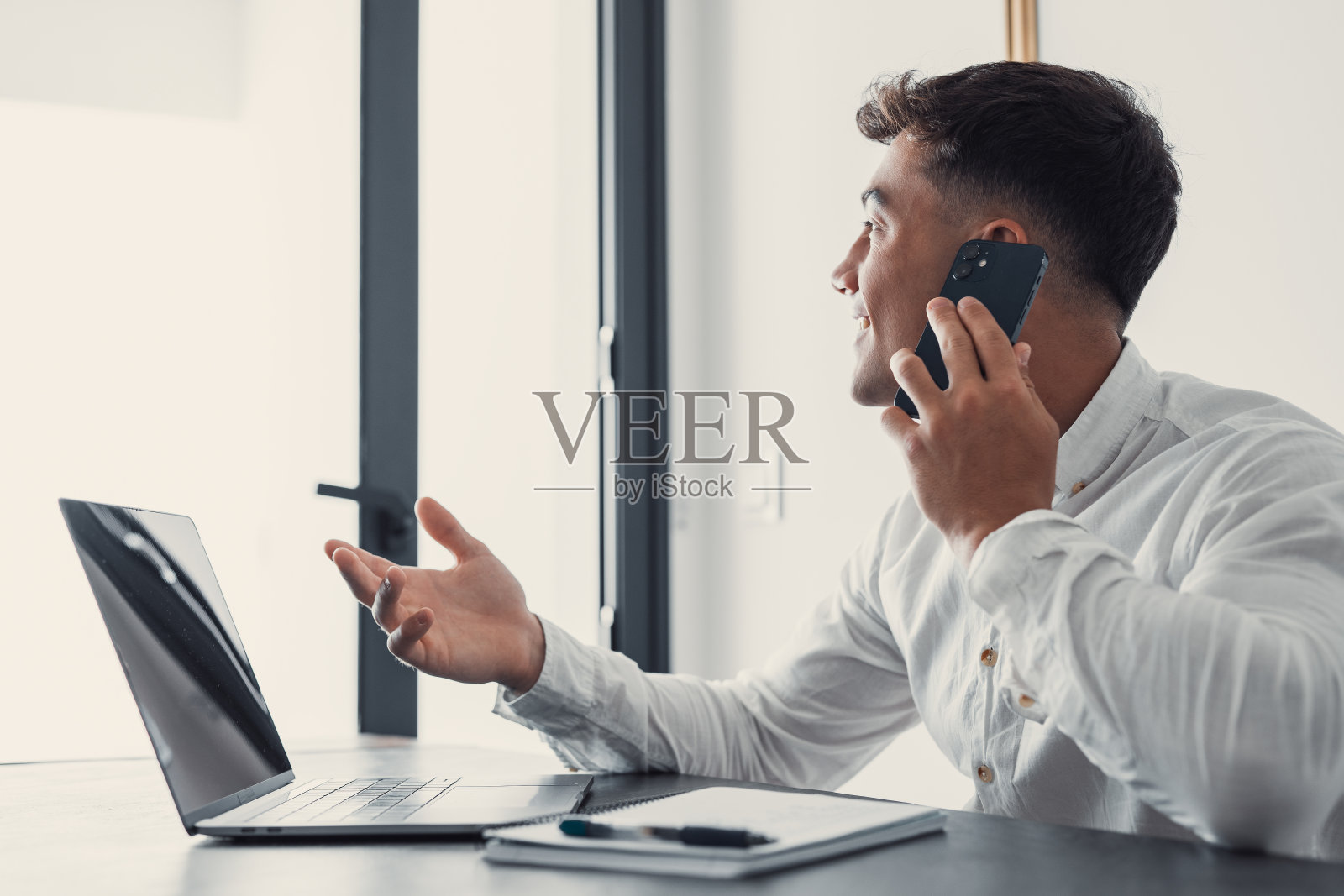 微笑的商人戴着眼镜打电话,坐在办公桌前用笔记本电脑,友好的经理通过电话咨询客户,快乐的人与朋友聊天从工作中分心照片摄影图片