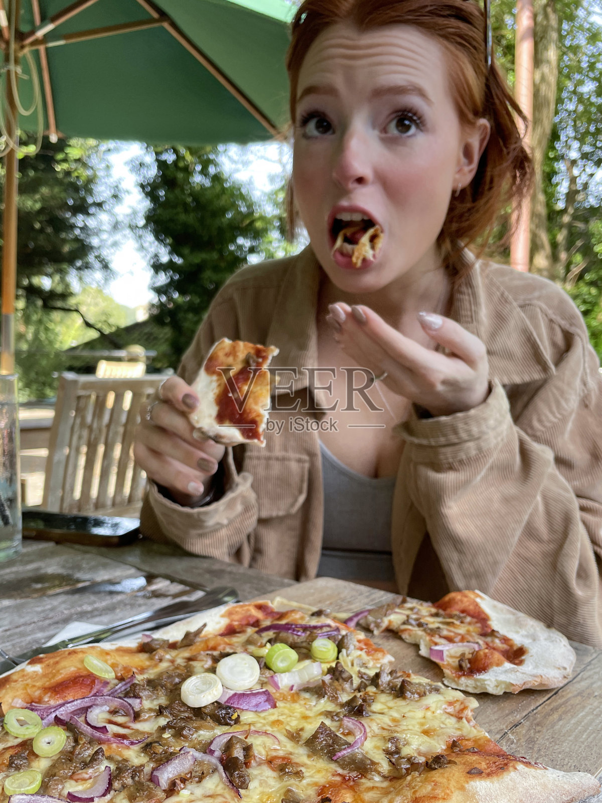 在餐馆的露天环境中,美丽的红头发年轻女子的特写图像,在一片新鲜出炉的烤酥鸭和海鲜酱披萨上燃烧的嘴,木制披萨桨,意大利和中国的融合美食,重点在前景照片摄影图片