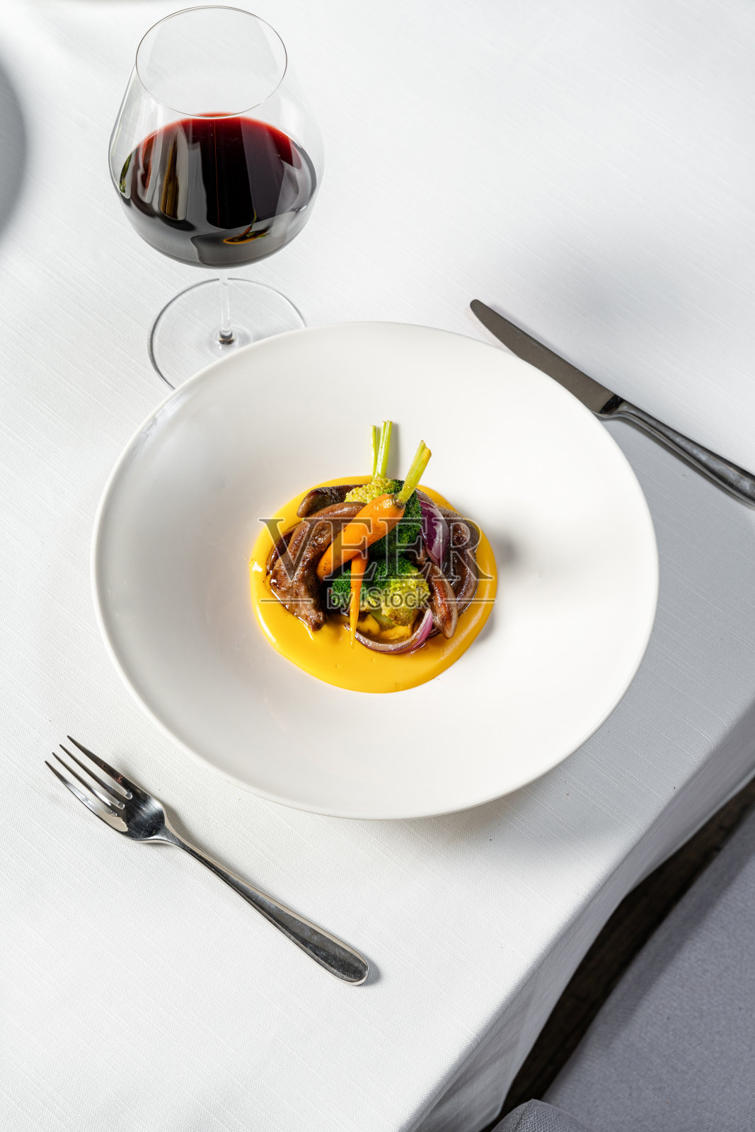用红酒炒肝,配上蒸蔬菜和南瓜泥,盛在白色宽边盘子里。盘子放在白色的桌布上,旁边是一杯红酒和餐具。照片摄影图片