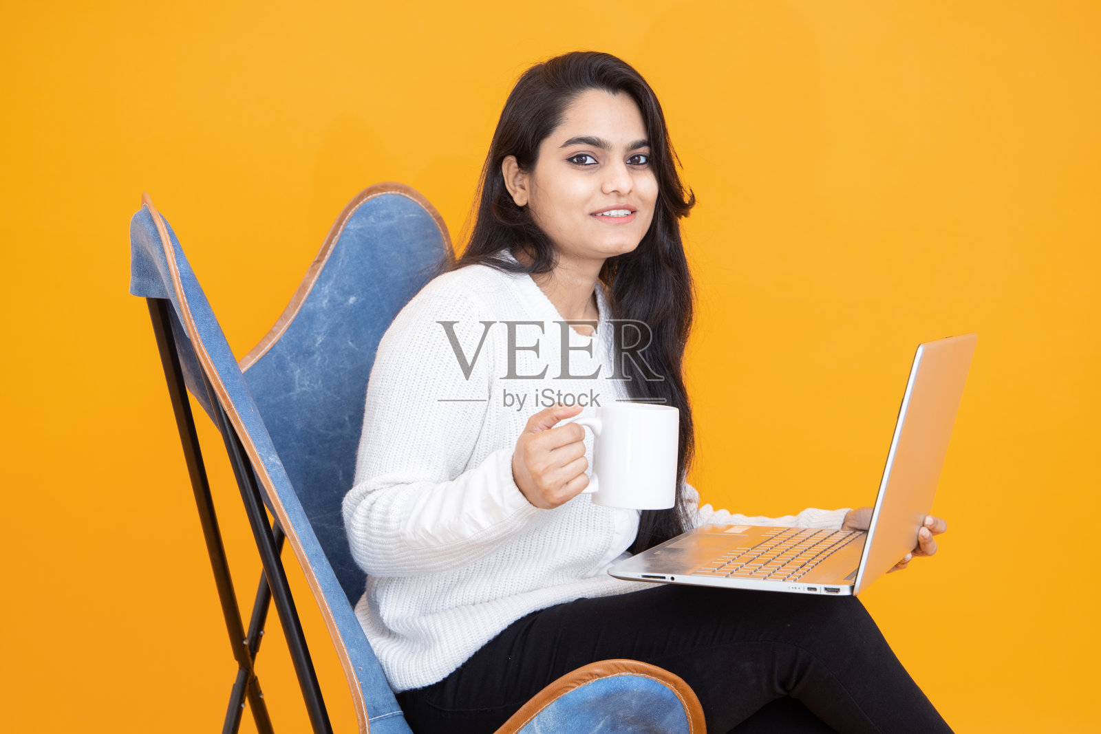 年轻的印度女孩拿着咖啡杯在笔记本电脑上工作,坐在椅子上,孤立的橘黄色背景。摄影棚拍摄,复印空间,亚洲女性使用电脑。技术的概念。照片摄影图片