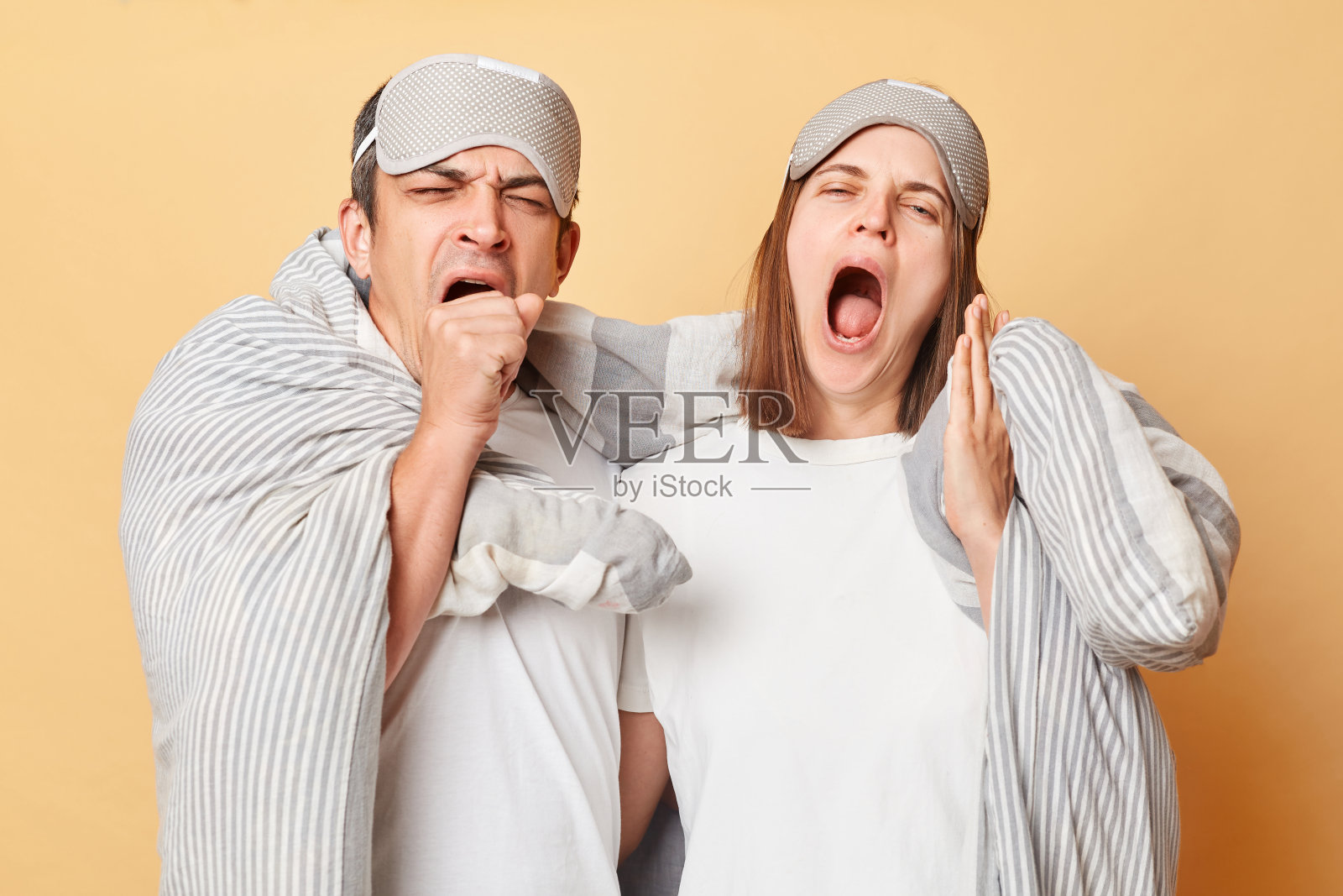 疲惫困倦的夫妇戴着眼罩,裹着毯子,在米色的背景下孤立起来,嘴巴张开,需要更多的时间小睡。照片摄影图片