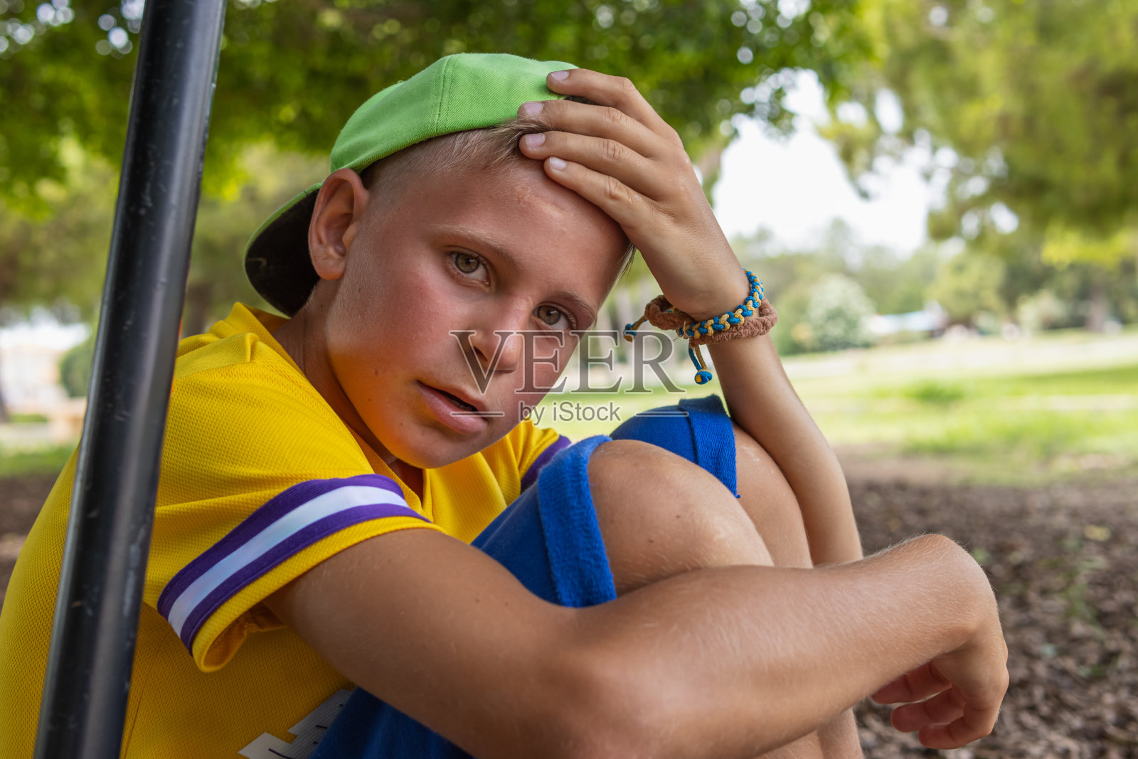 一个穿着黄色t恤,戴着绿色帽子的欧洲男孩坐在公园里,红着脸,一副疲惫的样子,孩子在新鲜的空气中玩着活跃的游戏照片摄影图片