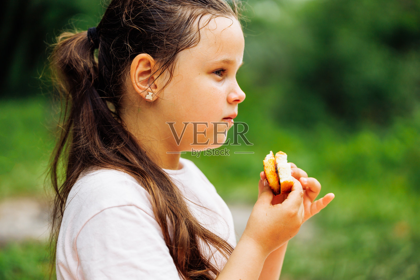 小女孩的侧视图,深色头发,拿着三明治,嚼着,坐在公园森林的绿树之间。照片摄影图片