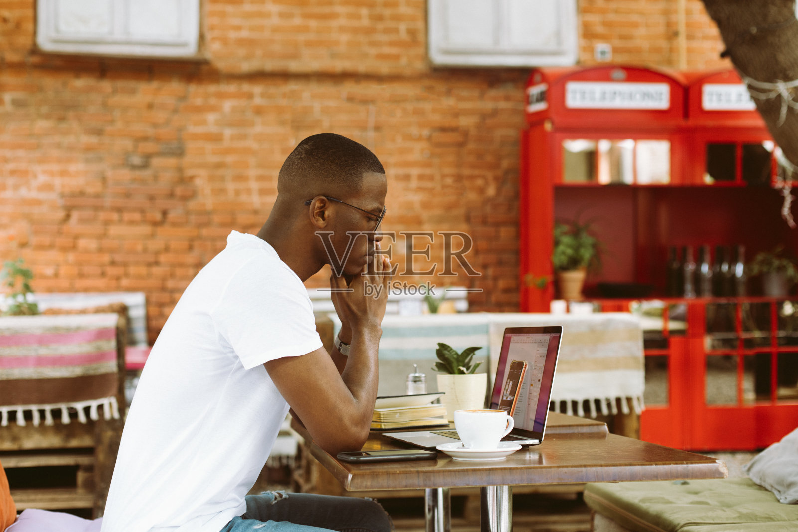 戴着眼镜的英俊的非洲裔美国人坐在夏日阳台咖啡馆的桌子旁,双手合十思考,解决问题。在笔记本电脑上在线工作,自由职业者,困难照片摄影图片