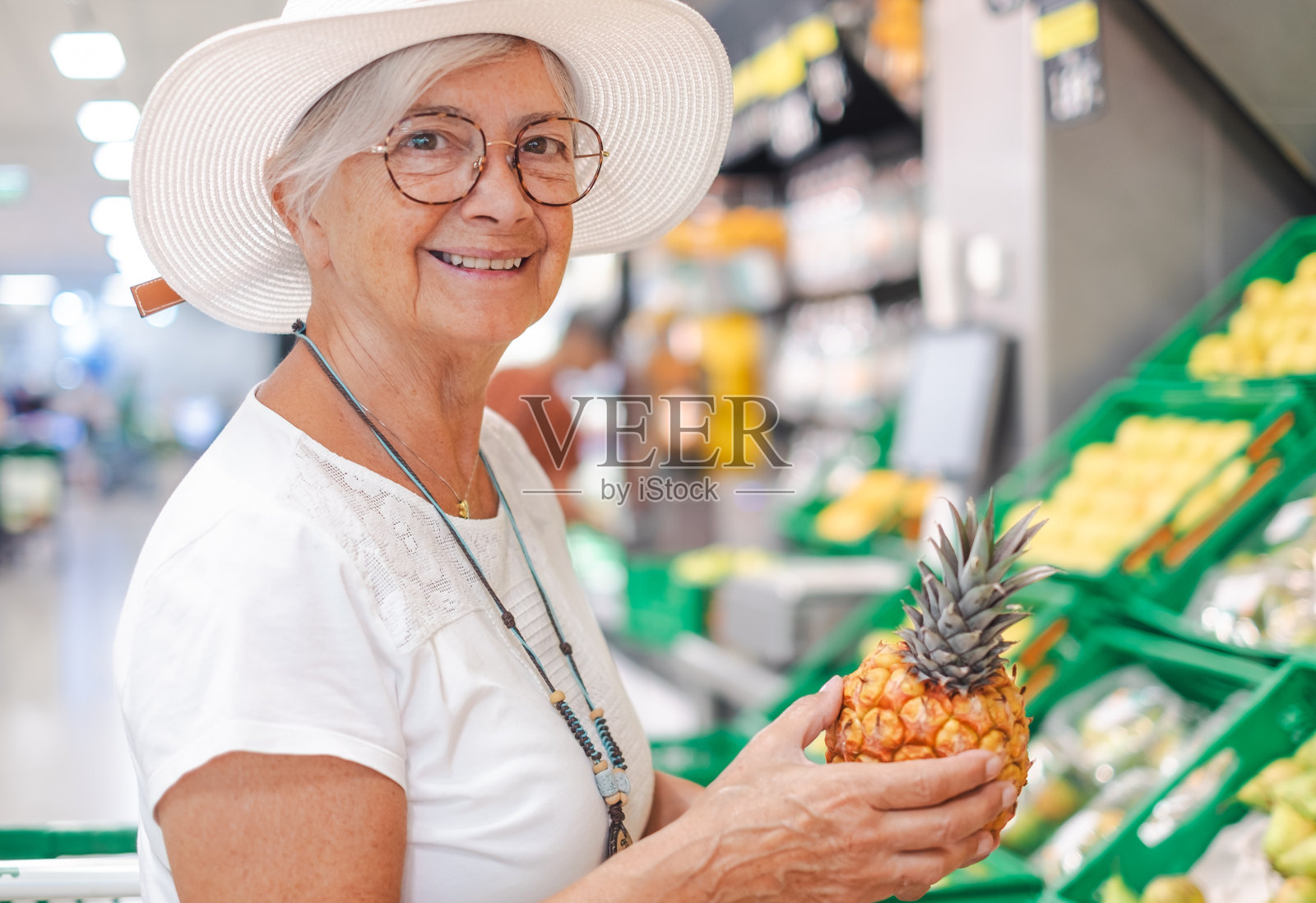 带着帽子微笑的老顾客女士在超市挑选新鲜水果,挑选成熟的菠萝。领取养老金的老人注意到物价的上涨照片摄影图片