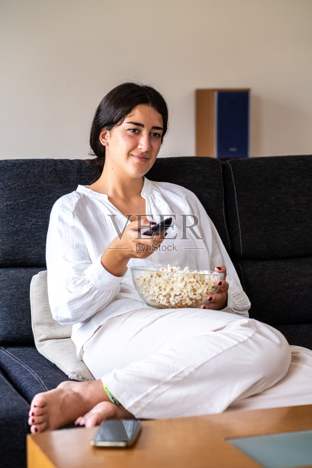 一位年轻女子准备好了爆米花,坐在客厅的沙发上用遥控器在电视上找节目。照片摄影图片