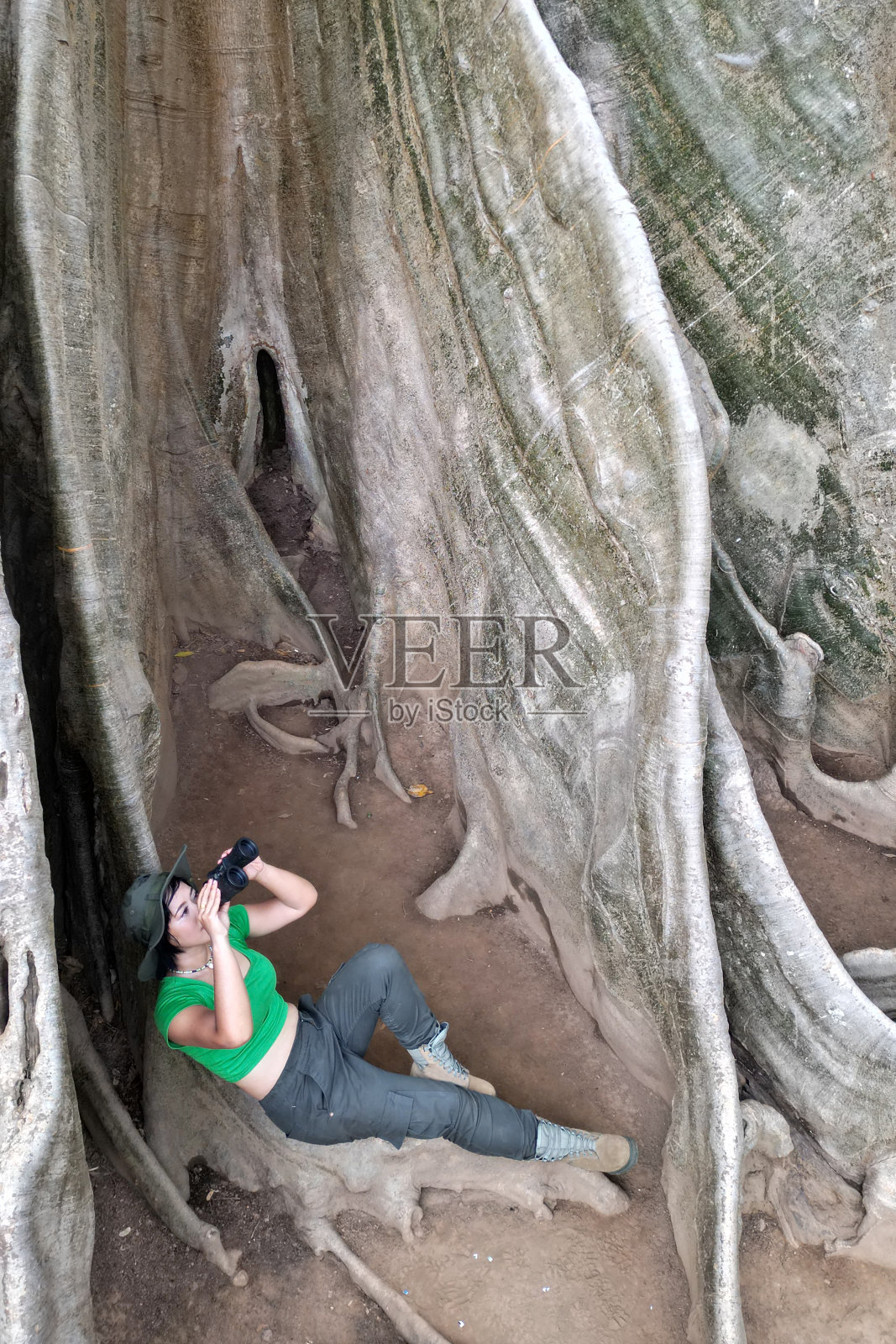 漂亮的亚洲女人用双筒望远镜看着巨大的树根。照片摄影图片