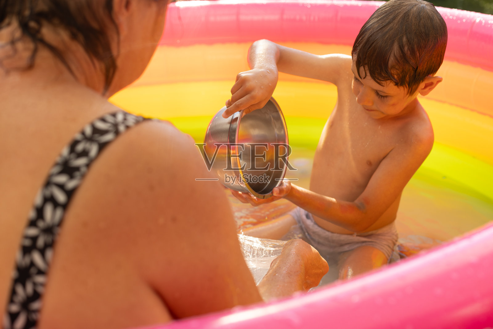 在一个阳光明媚的夏日,一个陌生的女人和一个男孩坐在室外的充气游泳池里,孩子在玩水。照片摄影图片