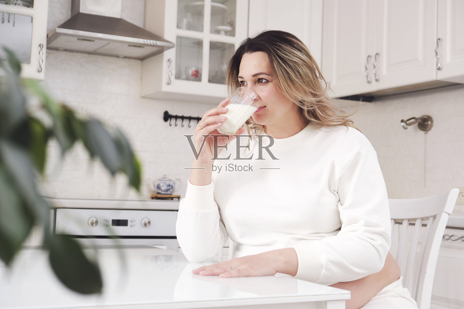 穿着白色套装的漂亮孕妇在明亮的厨房里喝牛奶。照片摄影图片