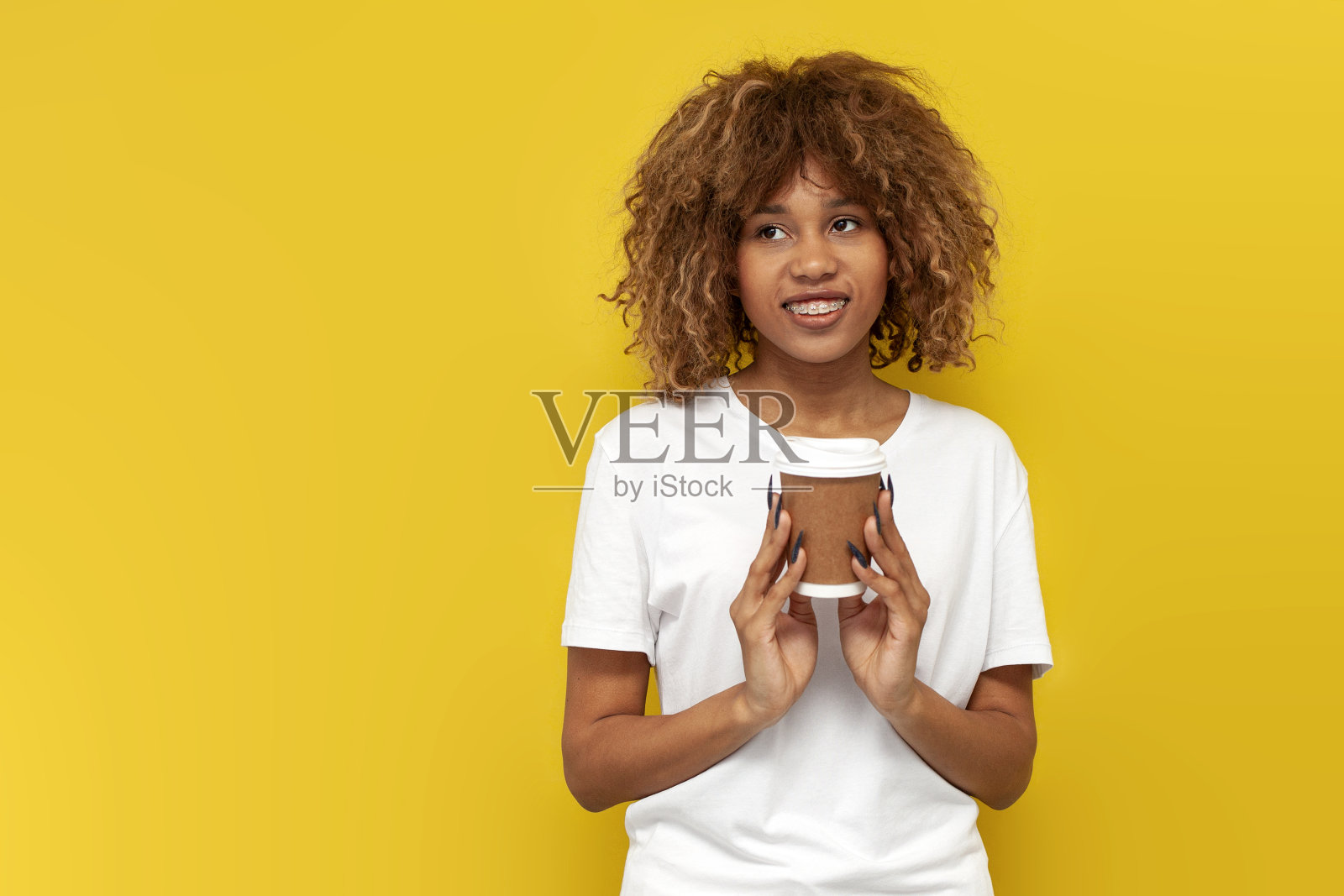 年轻卷曲的美国女孩戴着牙套,在黄色孤立的背景上拿着一杯纸咖啡照片摄影图片