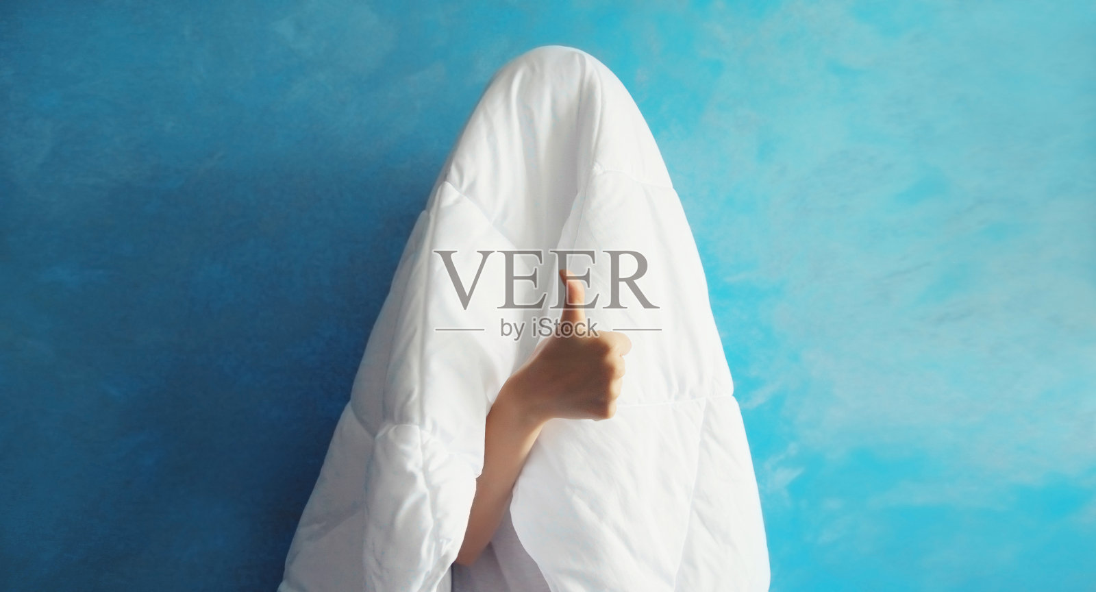 睡懒觉的女人睡醒后,用白毯子盖着头,在蓝色的背景上用手指做着凉爽的手势照片摄影图片