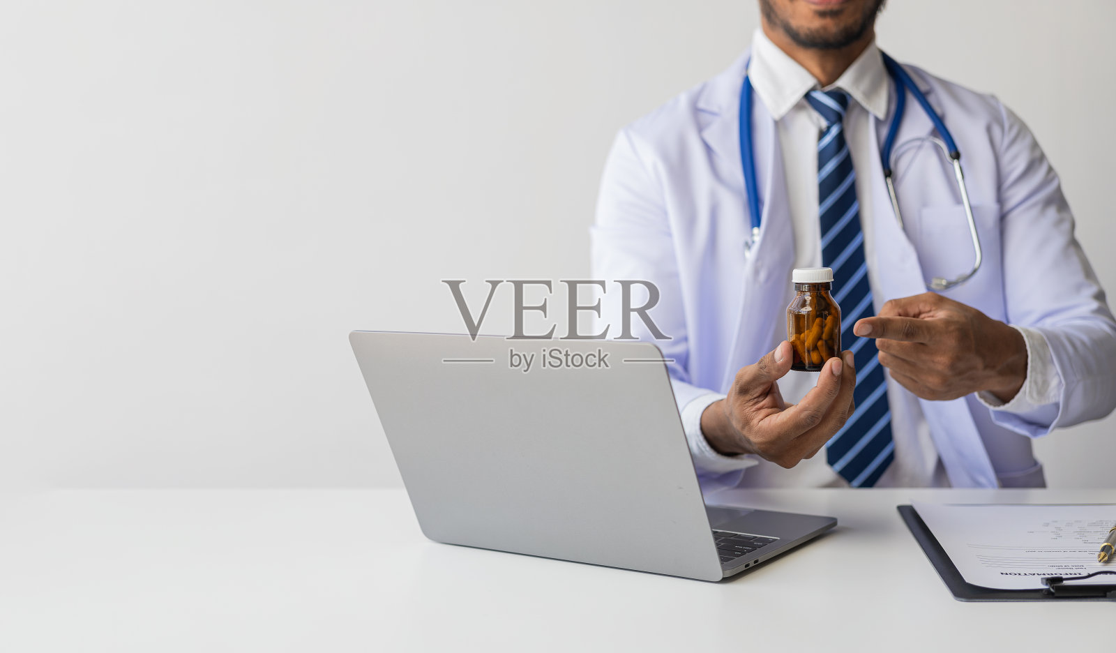 一个穿着白大褂拿着听诊器的男医生的画像。穿着白大褂的年轻非裔美国医生微笑着坐在医院办公室的桌子旁。照片摄影图片