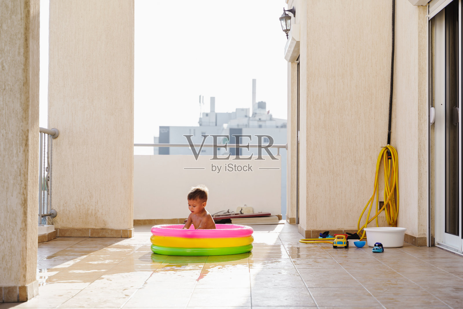 在炎热的夏日里,一个蹒跚学步的男孩在阳台上的充气圆形游泳池里欢快地玩耍照片摄影图片