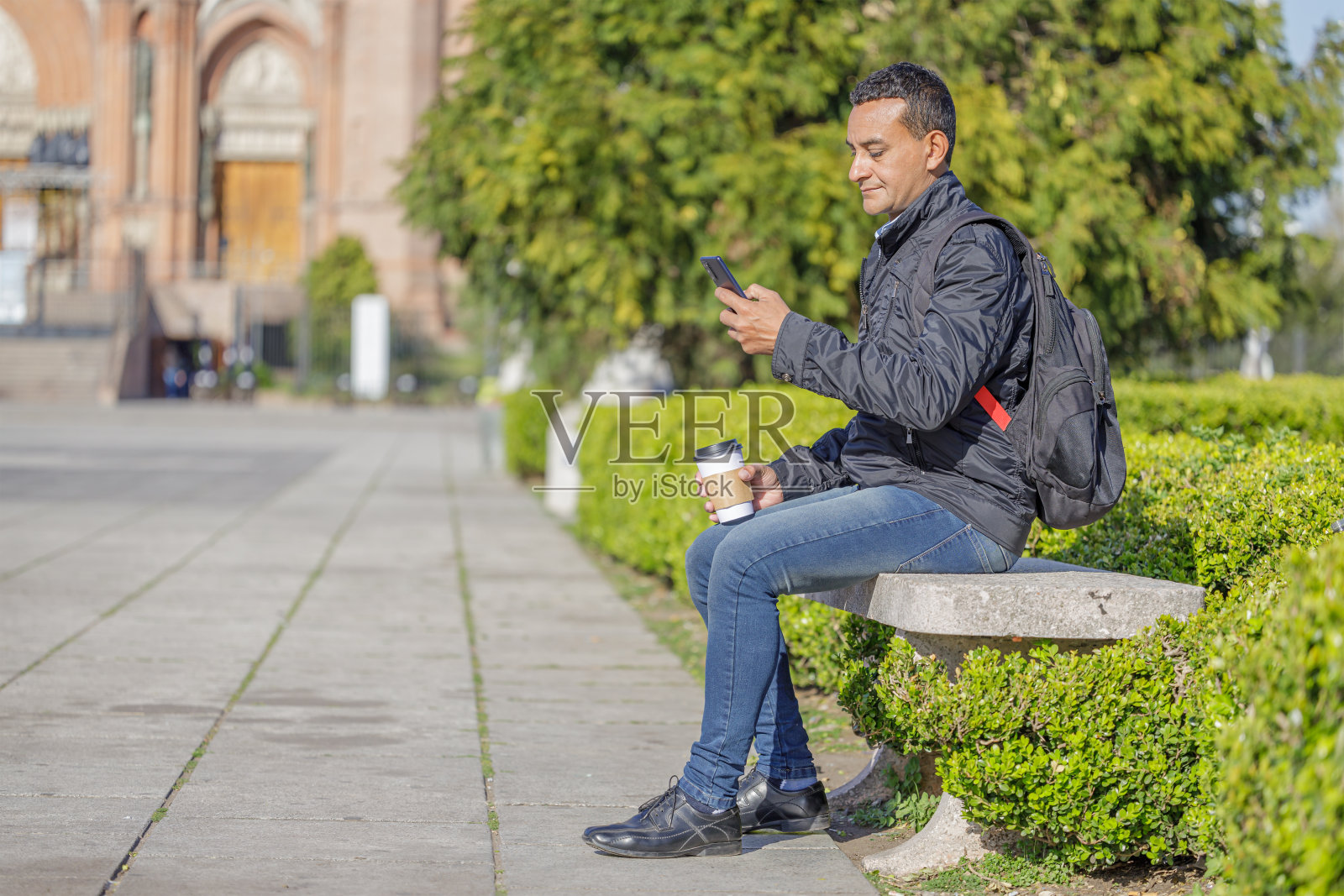 一个年轻的拉丁裔男子坐在公园的长椅上一边玩手机一边喝咖啡。照片摄影图片