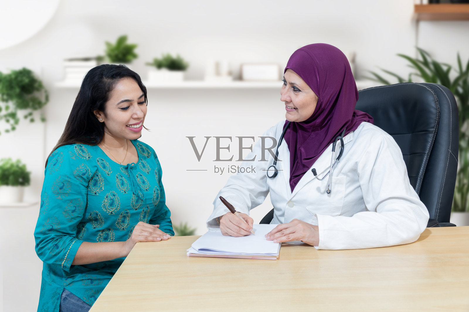 年轻的印度妇女在现代诊所咨询她对戴头巾的女穆斯林医生的问题。照片摄影图片