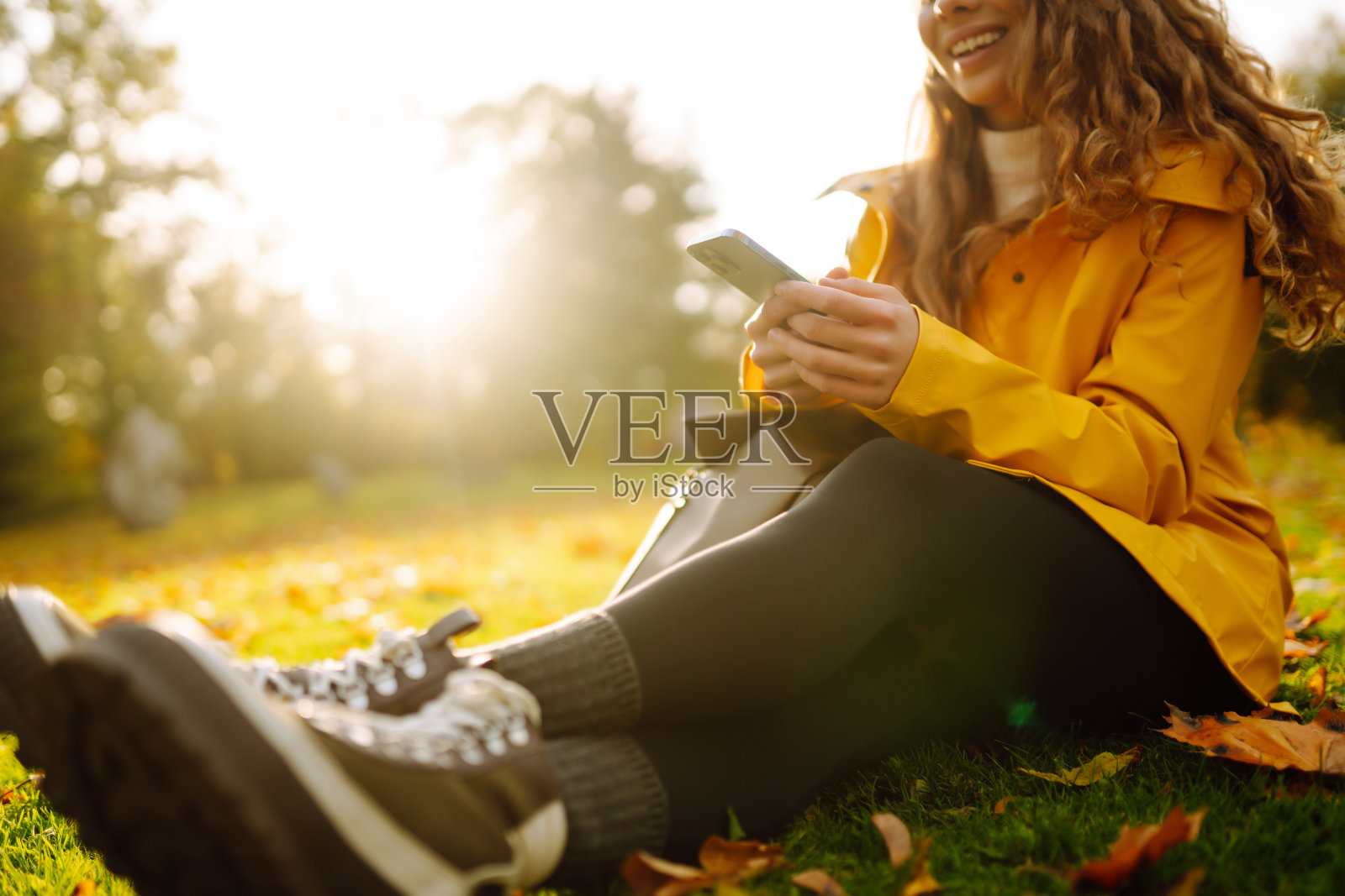 一个面带微笑的女人拿着电话坐在室外的草地上,周围是秋天的黄叶。照片摄影图片