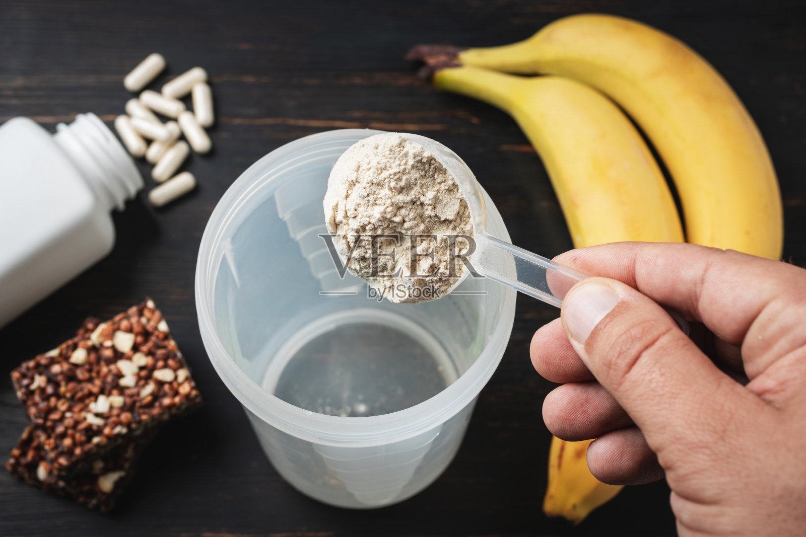 男手放入摇壶舀取部分乳清、大豆蛋白粉。白色的氨基酸胶囊,维生素,蛋白质巧克力棒和香蕉在一个黑色的木板上,健身食品补充剂照片摄影图片