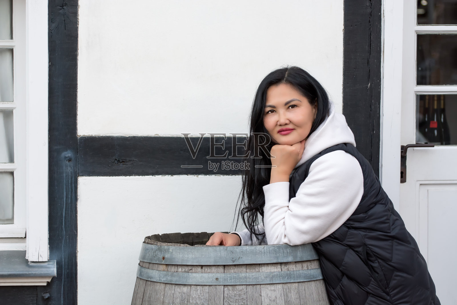 一个美丽的亚洲女人的街头肖像,她有着黑色的长发,胳膊肘倚在一个木制的啤酒桶上,背景是德国一座半木结构的老房子。布里亚特妇女穿着黑白相间的衣服。照片摄影图片
