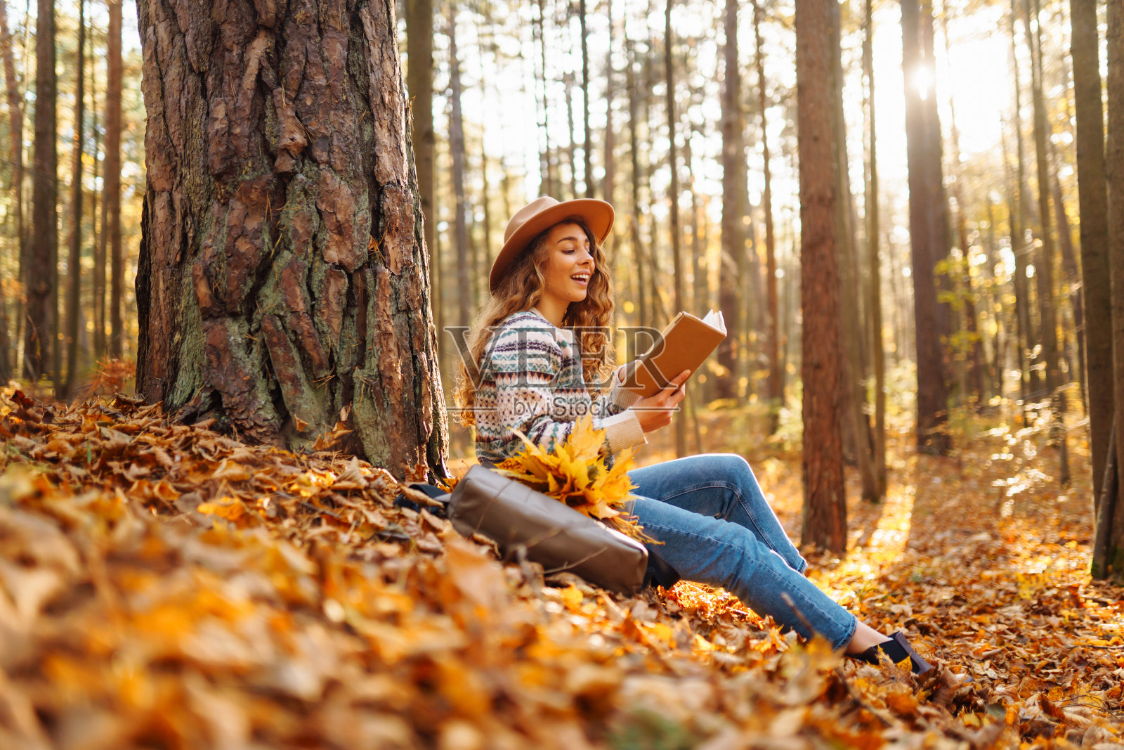 一个戴着帽子的女游客坐在秋天的森林里的黄叶上看书。照片摄影图片