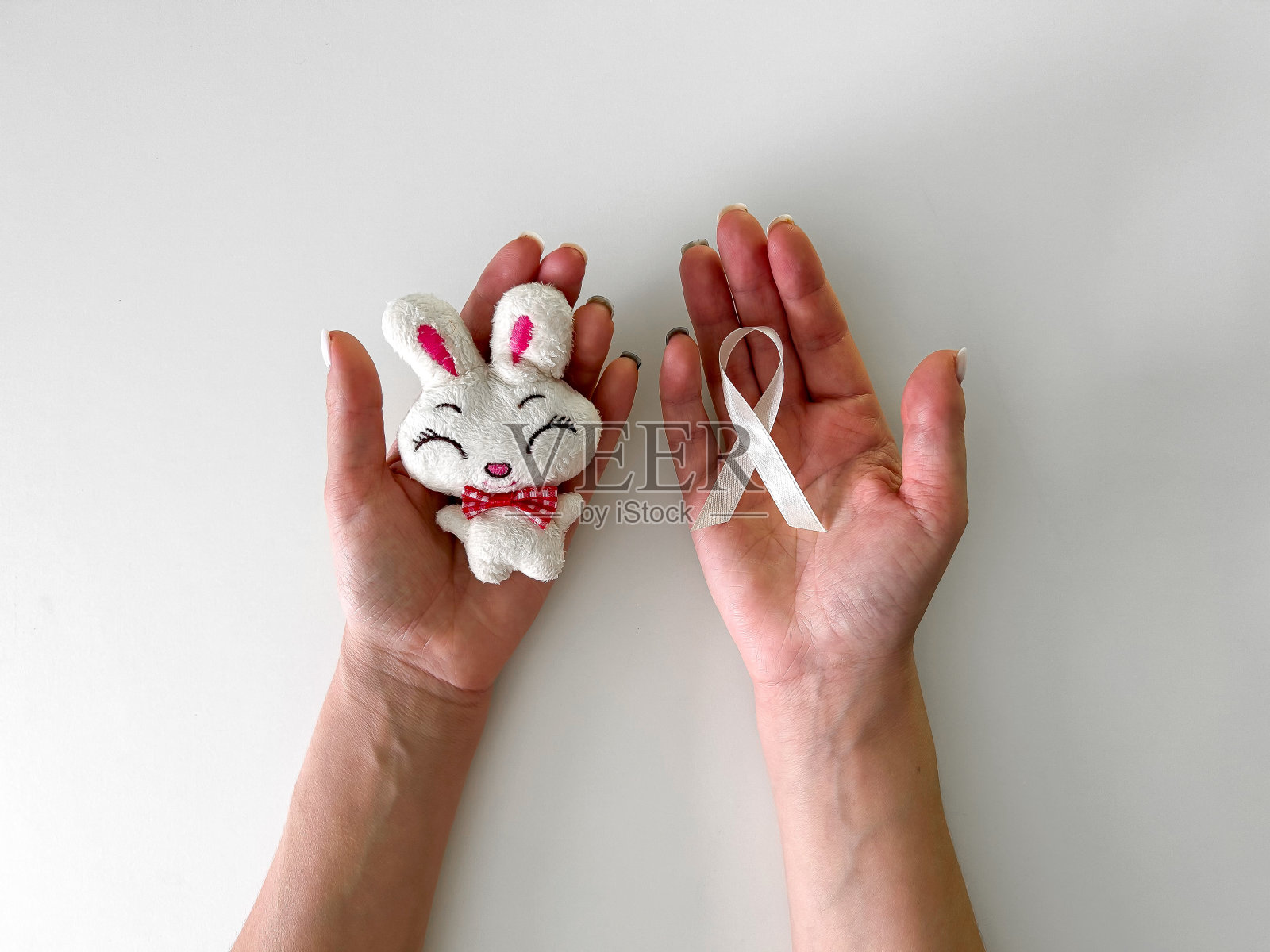 一个女孩的手拿着一个儿童兔子玩具和一条白色的丝带,以纪念儿童癌症意识,在白色的背景上。儿童癌症日,2月15日。照片摄影图片