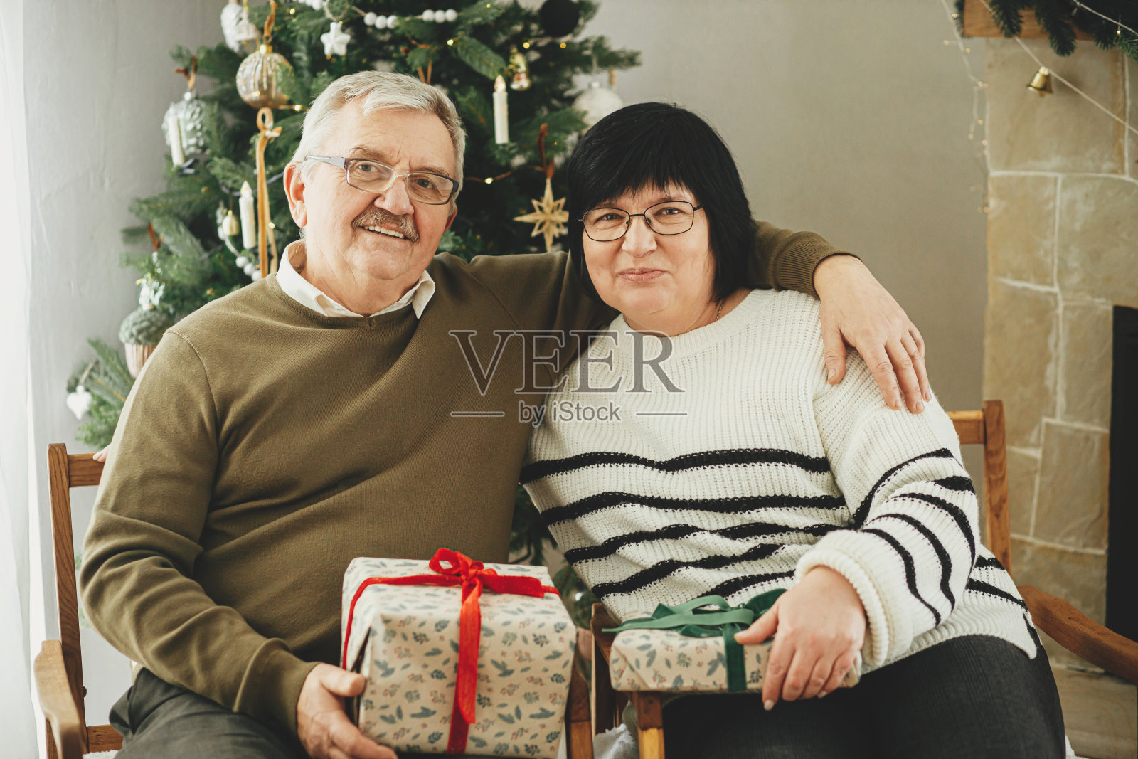 美丽的老夫妇交换圣诞礼物,坐在温暖的壁炉在时尚的节日装饰的客厅。快乐的老年家庭享受舒适的夜晚和礼物。节日快乐照片摄影图片