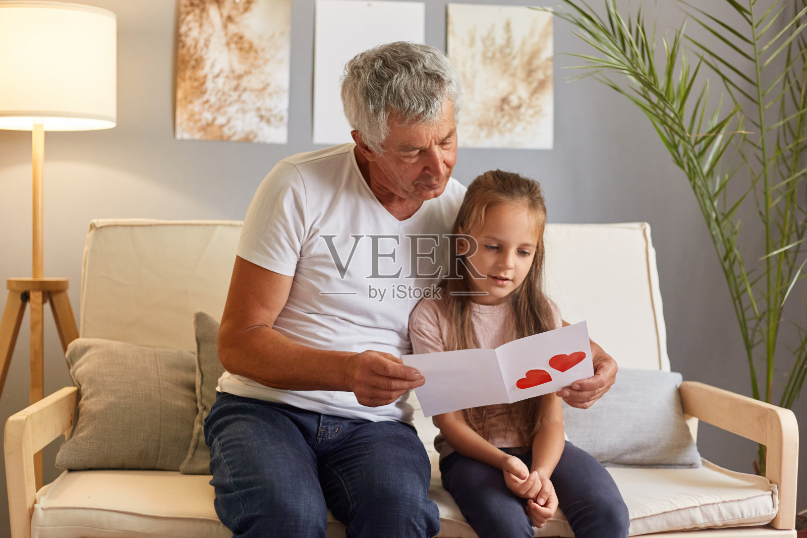 爷爷和孙女坐在沙发上阅读手工制作的红色心形贺卡,一起坐在家里的沙发上。照片摄影图片