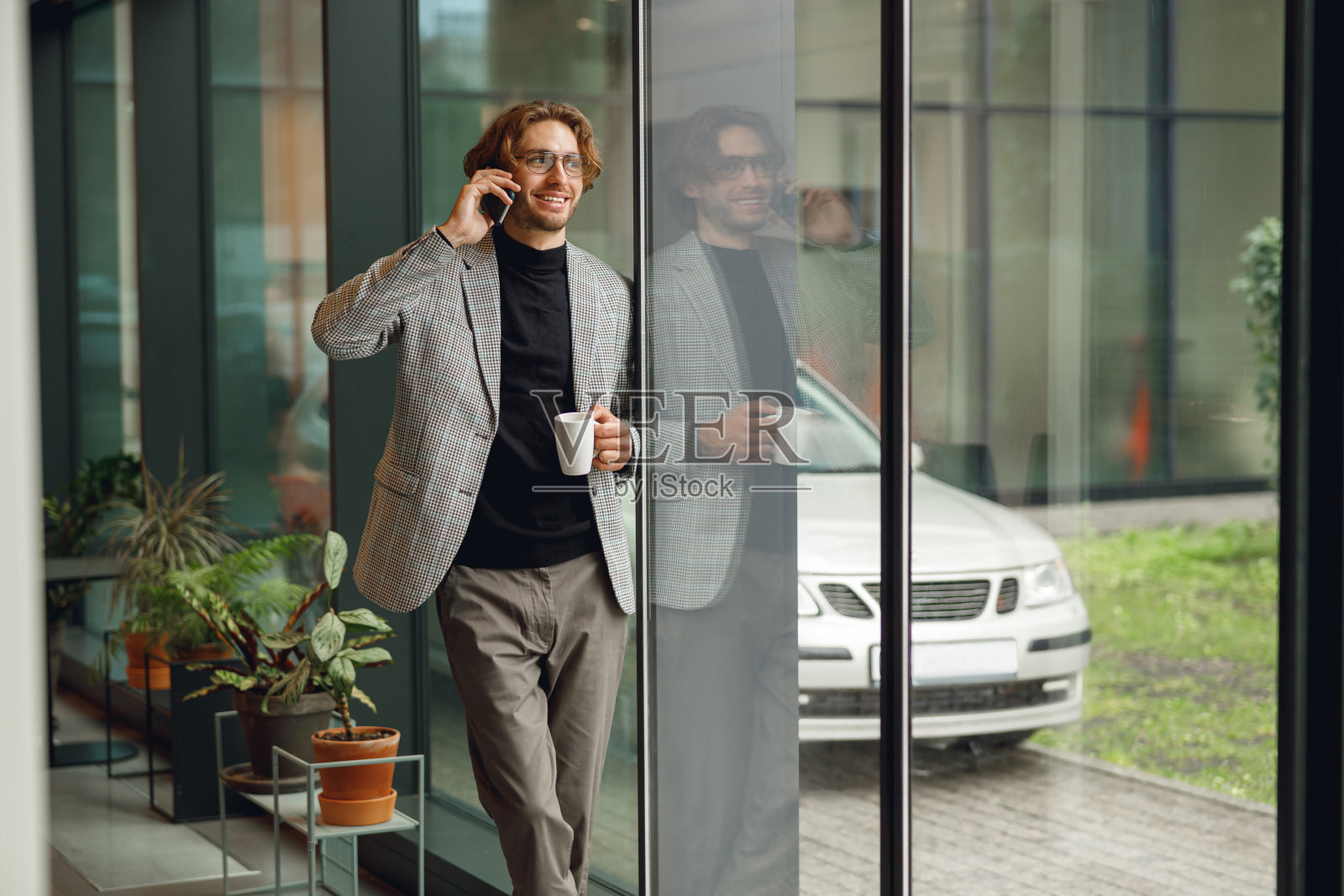 戴着眼镜的商人站在靠近窗户的现代化办公室里讲电话照片摄影图片