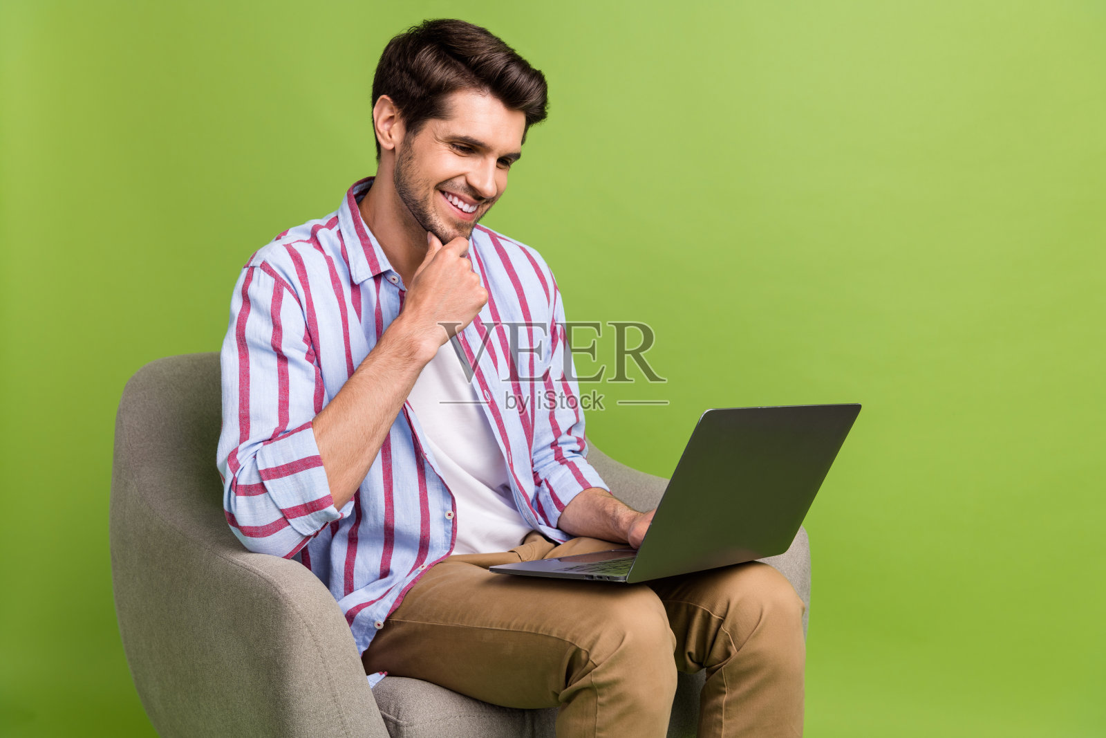 照片积极聪明的男人留着胡茬,穿着条纹衬衫,看着笔记本电脑思考,坐在扶手椅上孤立的绿色背景照片摄影图片