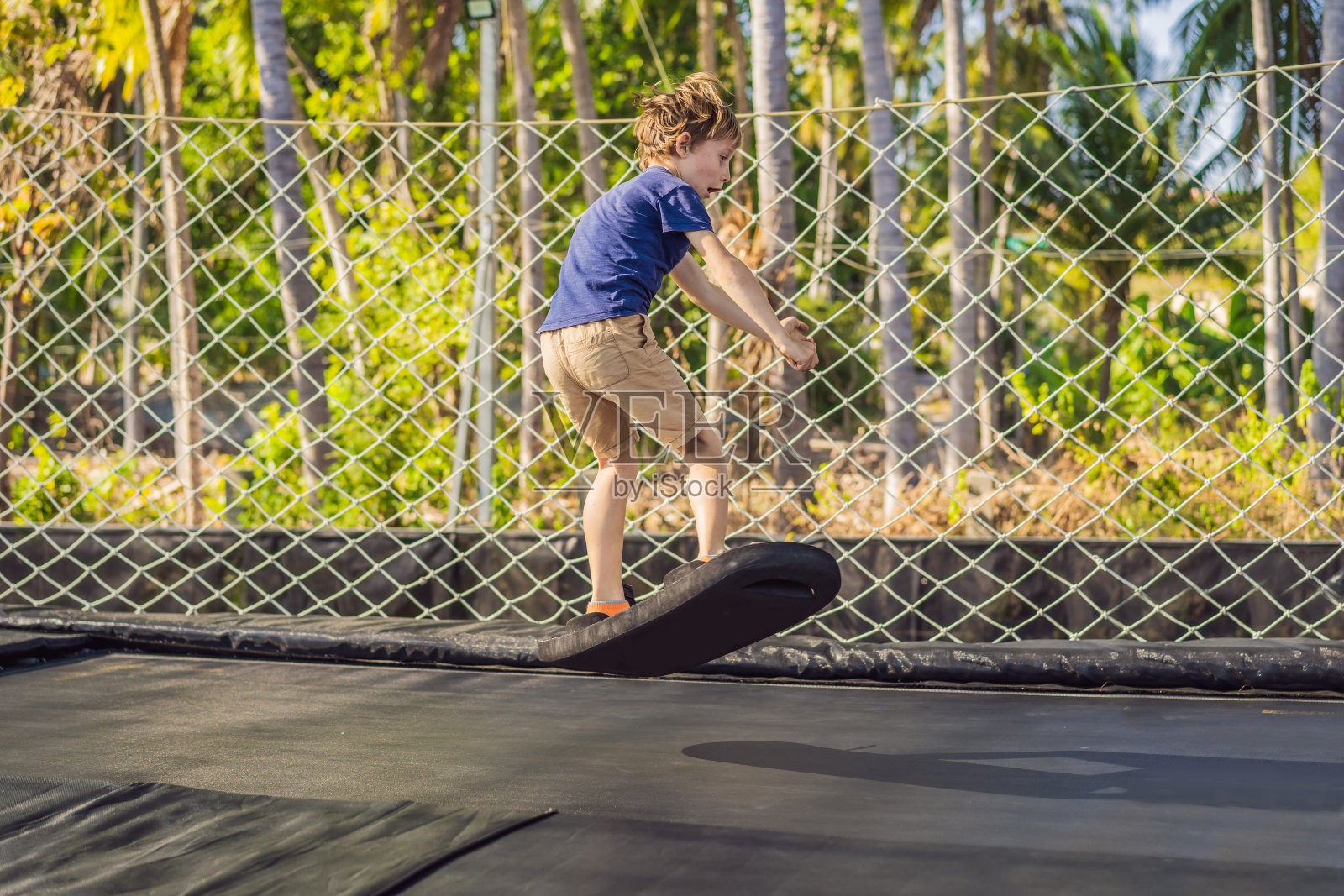 快乐的男孩在一个柔软的蹦床板上跳跃在室外的蹦床上,背景是棕榈树。蹦床板就像滑水板、滑板或滑雪板教练,可以磨练你的跳跃技能照片摄影图片