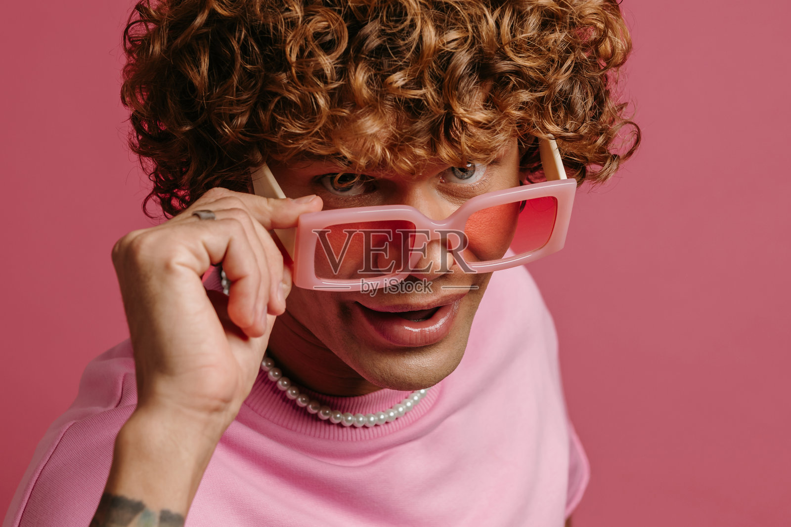 酷酷的年轻卷发男子在复古风格的眼镜看着相机在粉红色的背景照片摄影图片