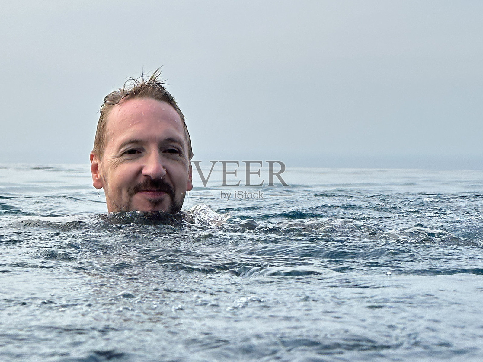 白人男子在无边泳池里游蛙泳的特写图像,溅起水花,阴天背景,聚焦于前景照片摄影图片